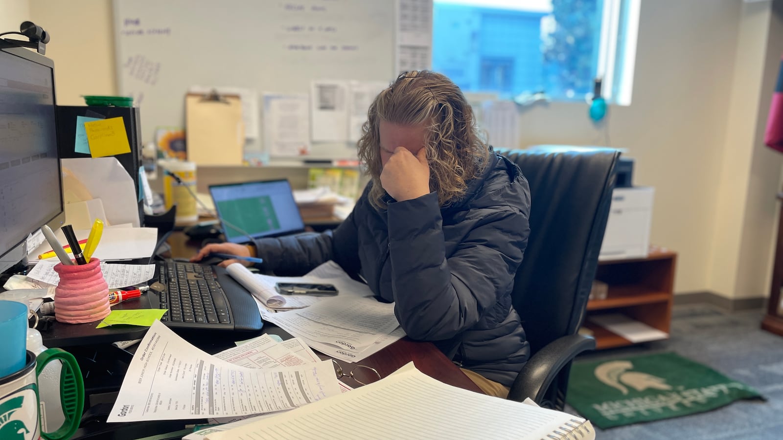 A nutrition services supervisor places her hand over her face in exasperation behind a desk piled with notebooks and paper.