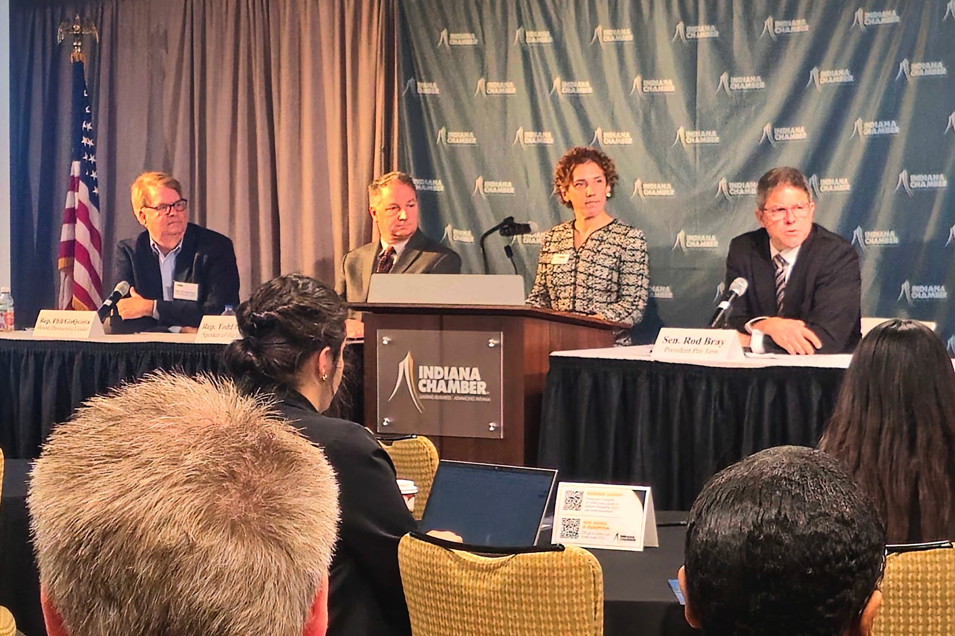 Three men and a woman in business attire sit in front of a group of people.