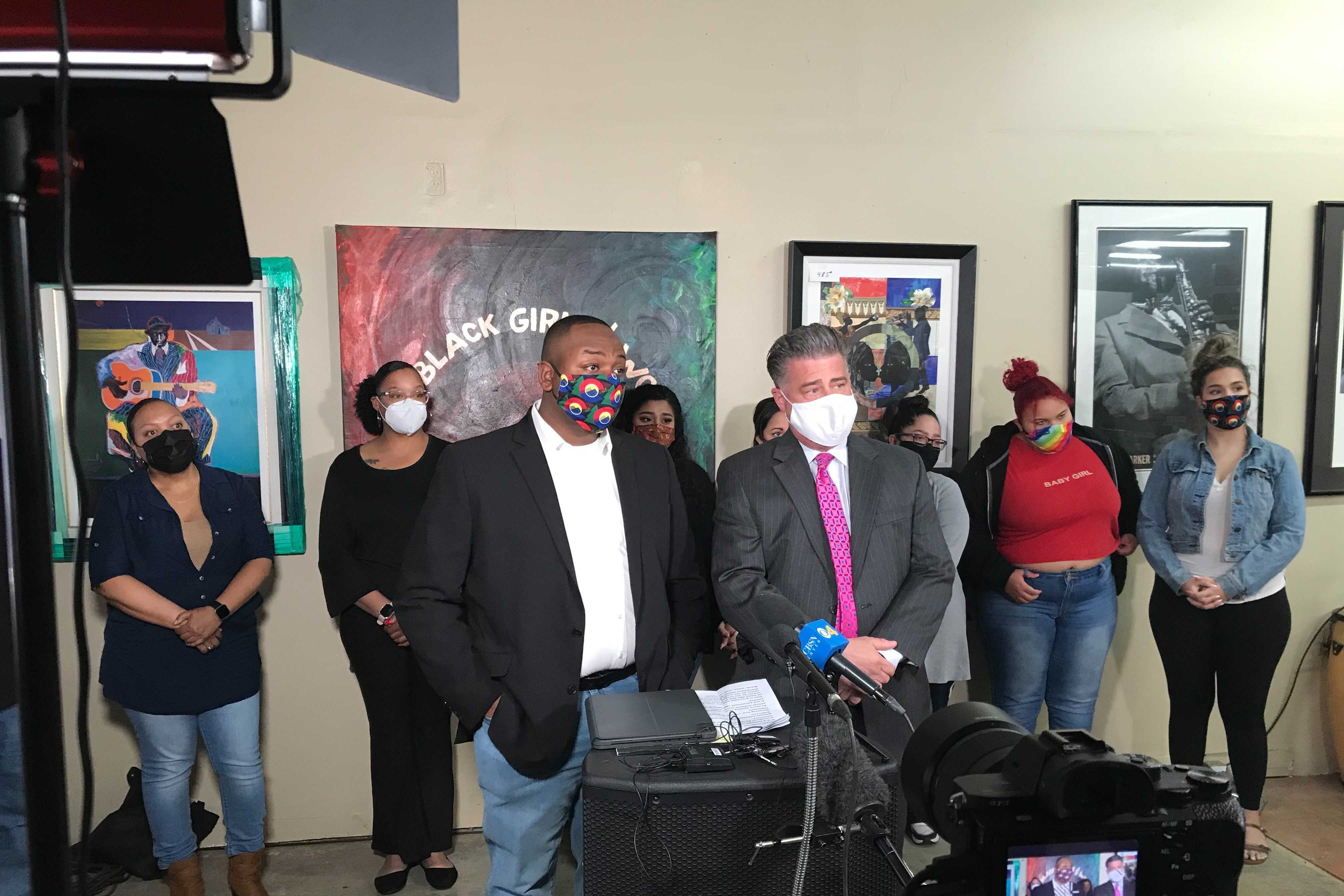 Denver school board member Tay Anderson, left, and his attorney Christopher Decker stand at a podium at a news conference. Supporters stand behind them.