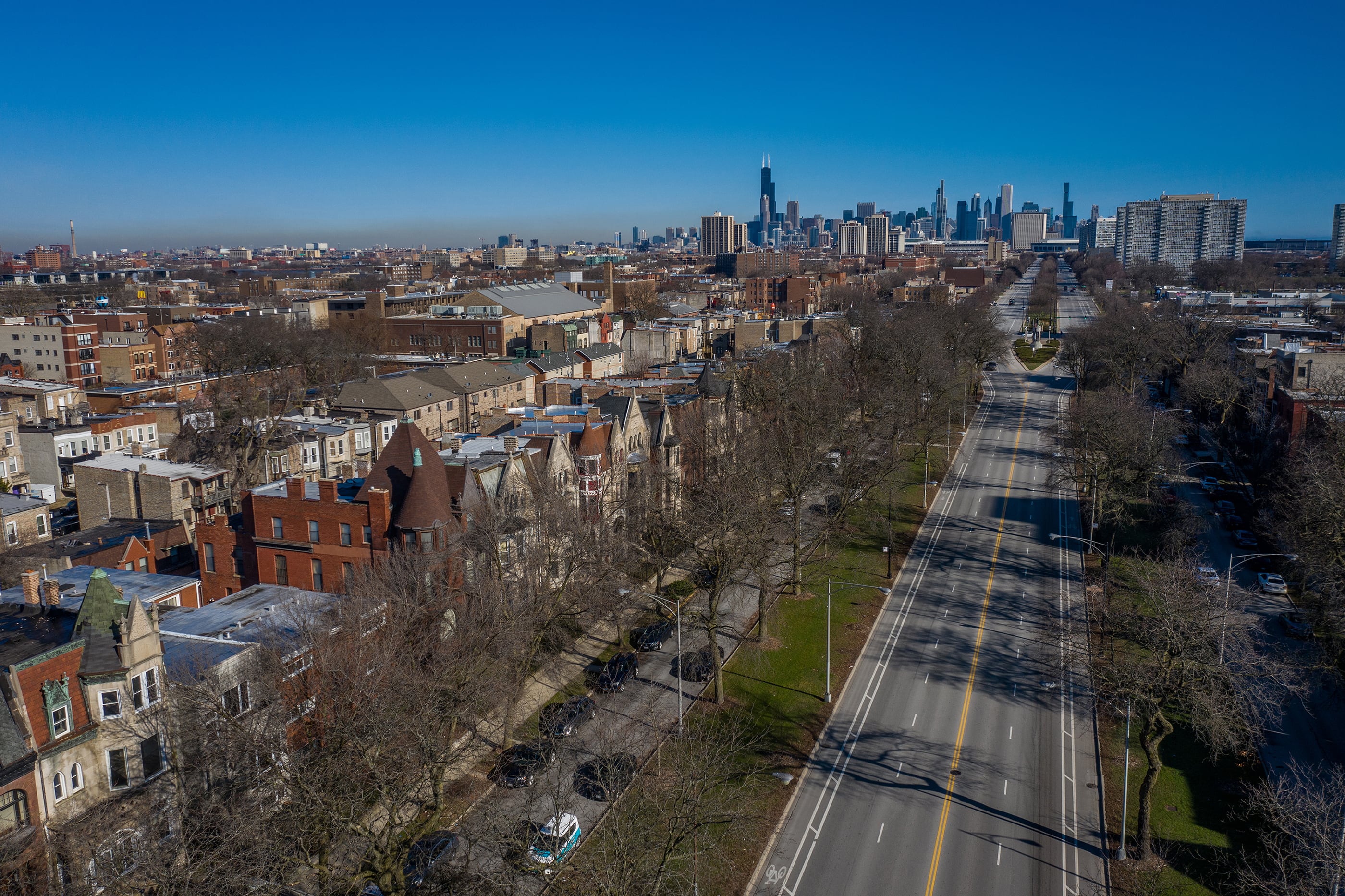An aerial photograph of the Chicago skyline in the background with a large four lane road that stretches all the way through the whole photo and rows of buildings and houses on the left side.