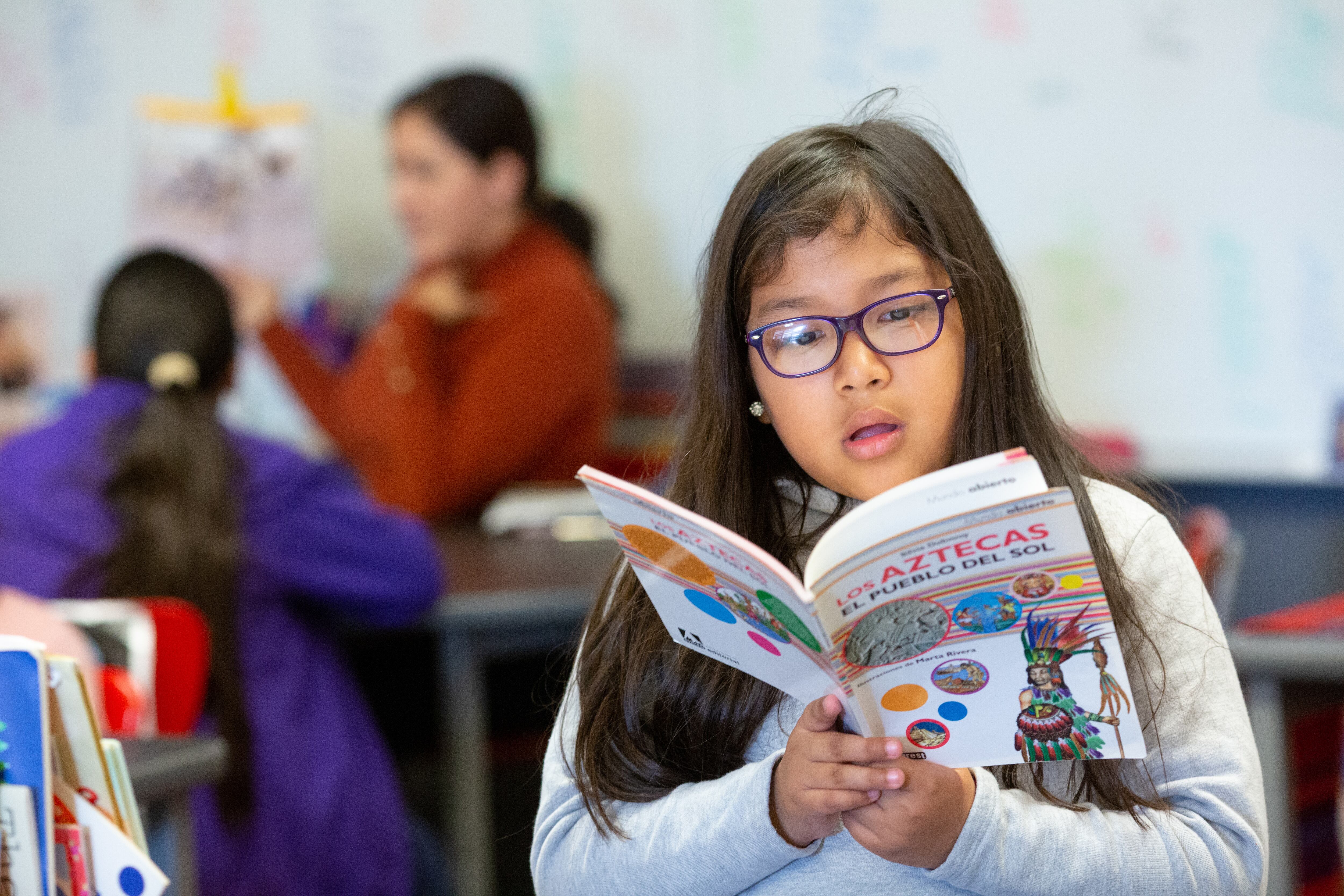 A girl wearing glasses reads a book in Spanish to  herself.