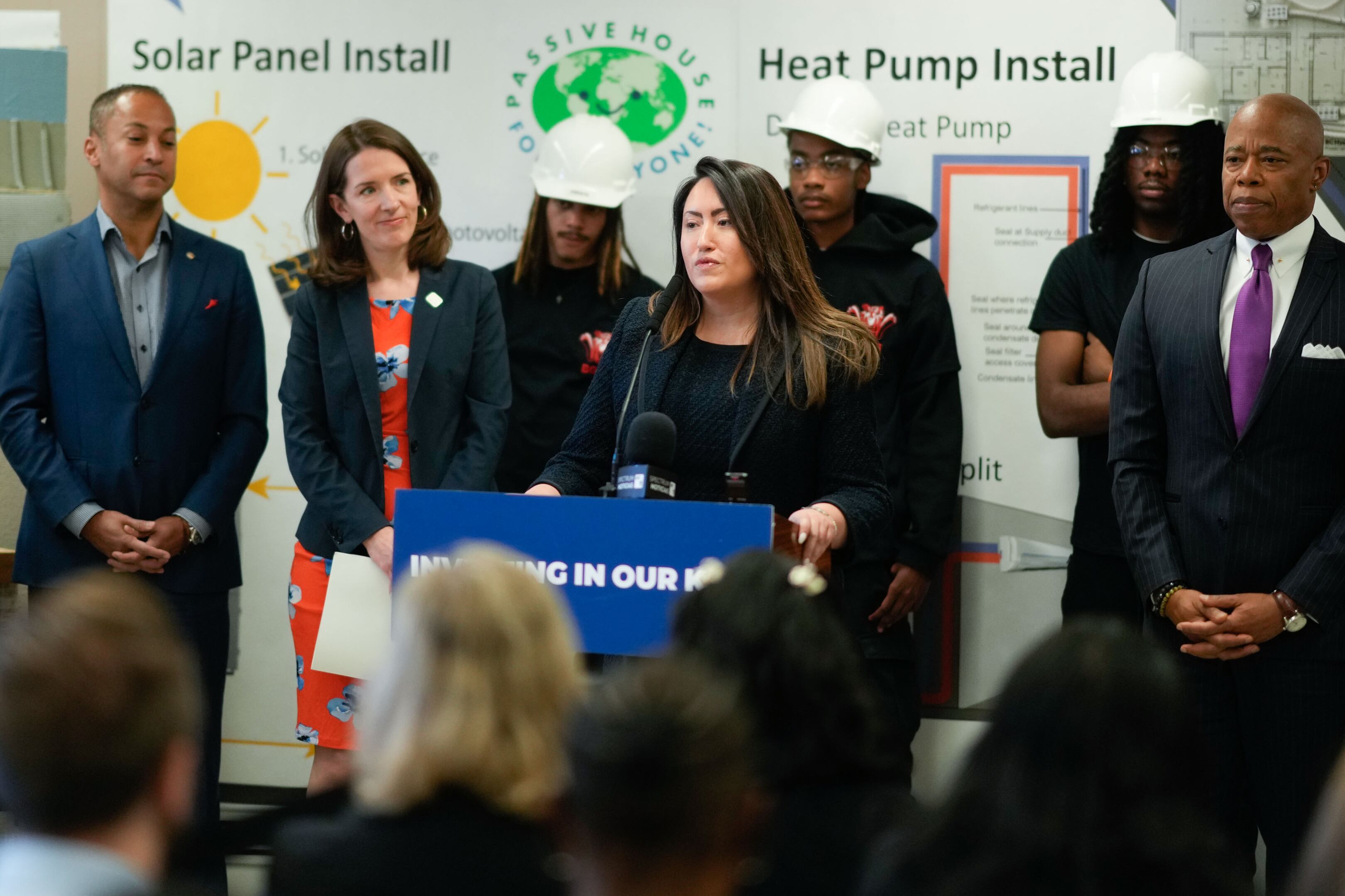 A woman in a dark suit speaks from behind a podium while a group of adults stand behind her.