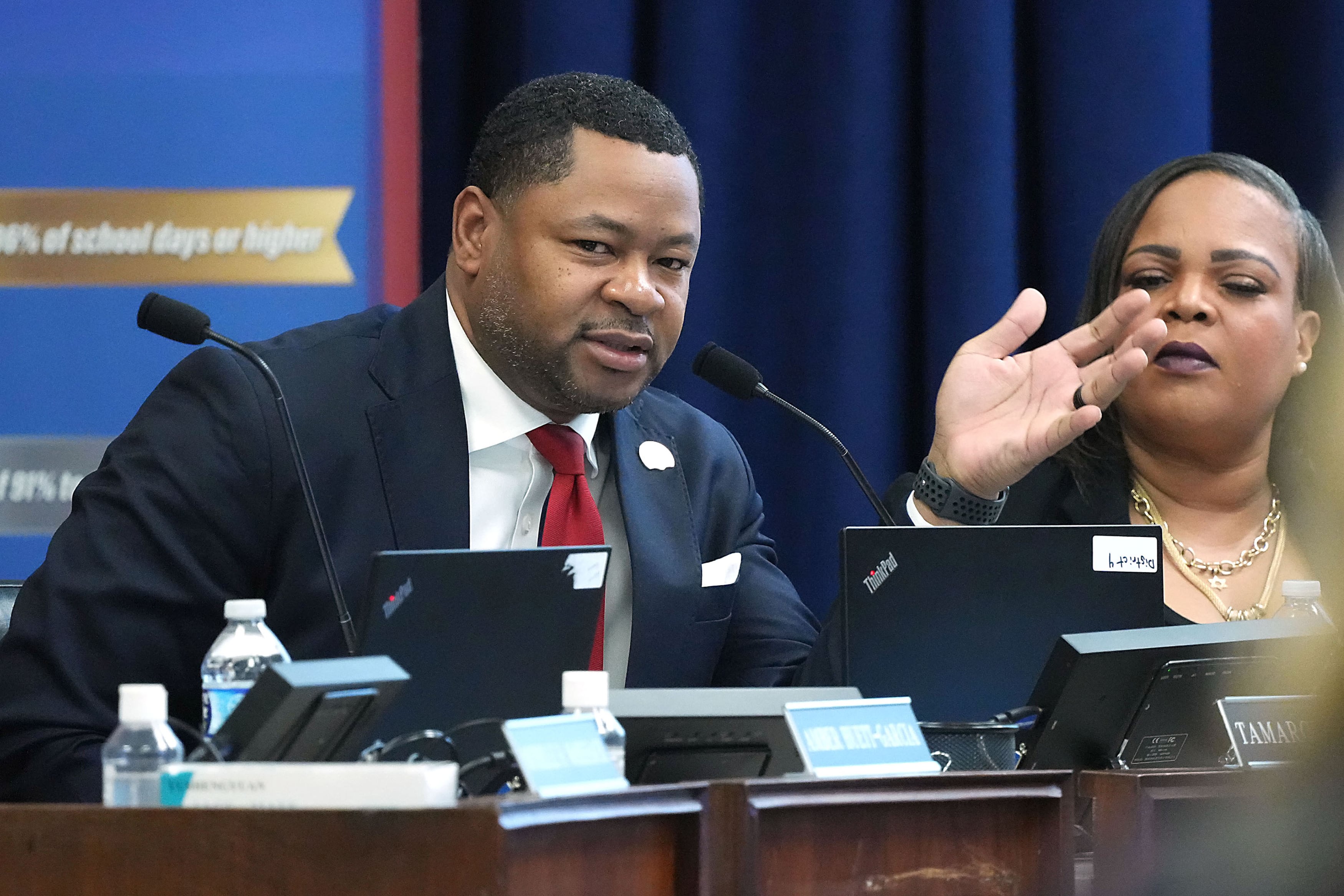 A photograph of a Black man and woman wearing business clothes sitting next to each other in a conference room and speaking into a microphone.