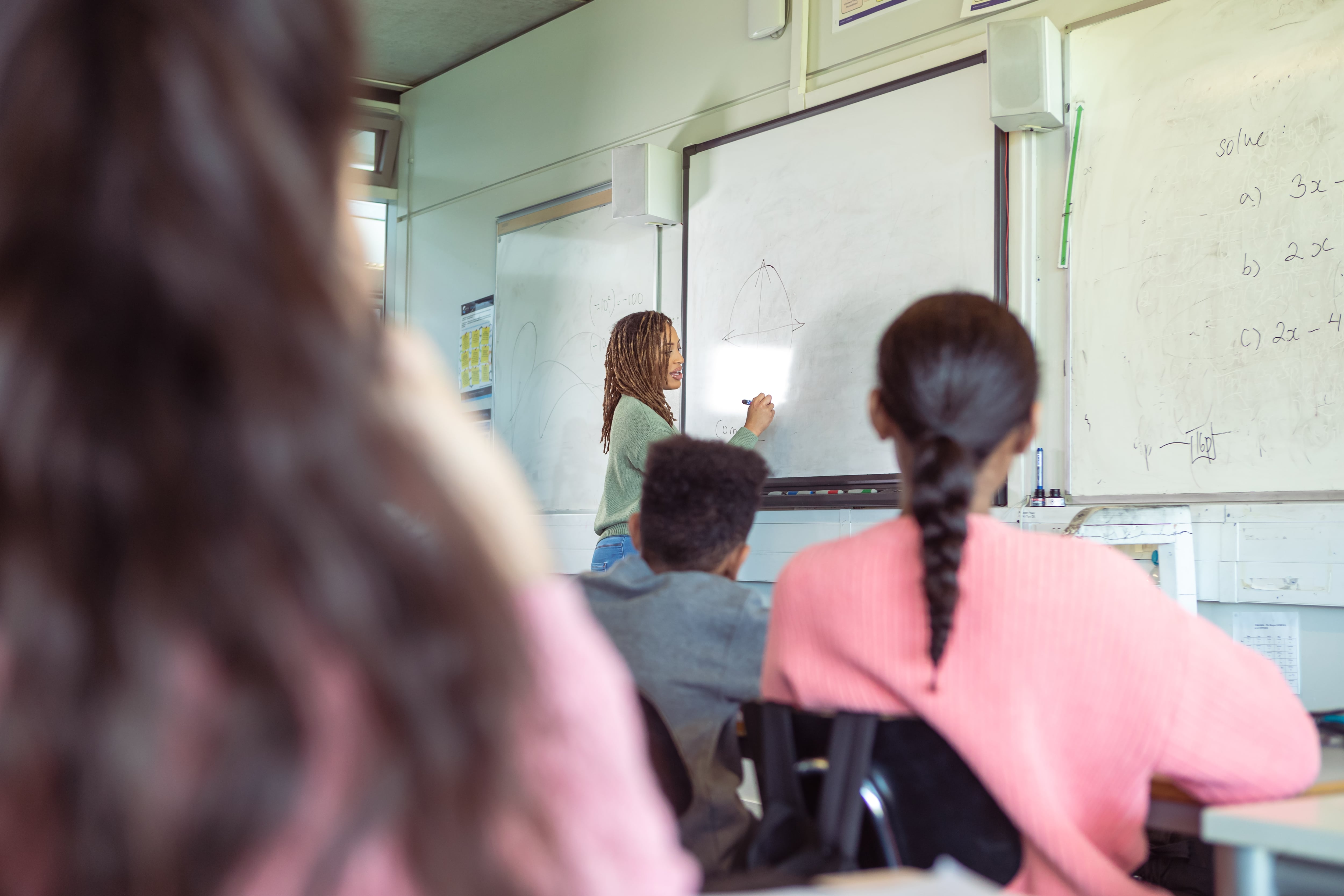 A cheerful black female teacher stands at the front of her classroom and writes on a whiteboard while instructing a multiracial group of elementary students who are attentively listening while seated at their desks.