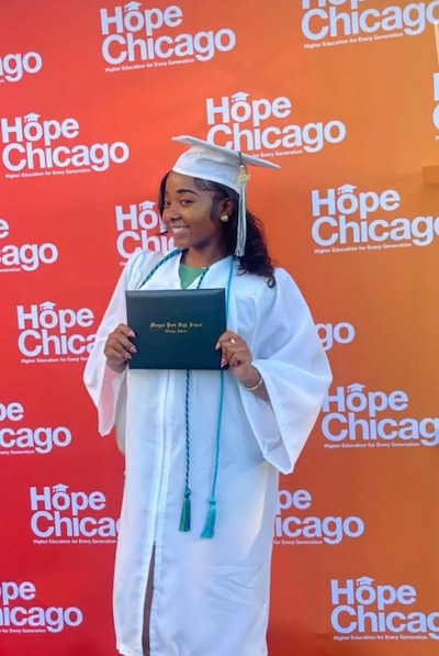 A high school senior with dark hair and wearing a white graduating cap and gown holds a diploma while posing in front of a red and orange banner that has white words that reads "Hope Chicago".