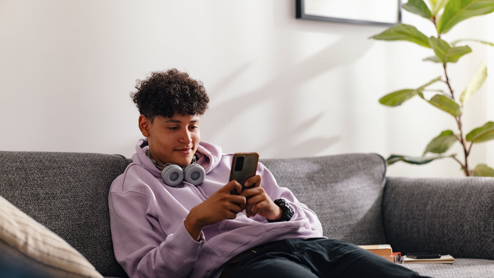 A teenage boy in a purple hoodie and headphones sits on a sofa, using his smartphone in a bright, cozy living room. The scene evokes a sense of modern adolescence and connectivity.