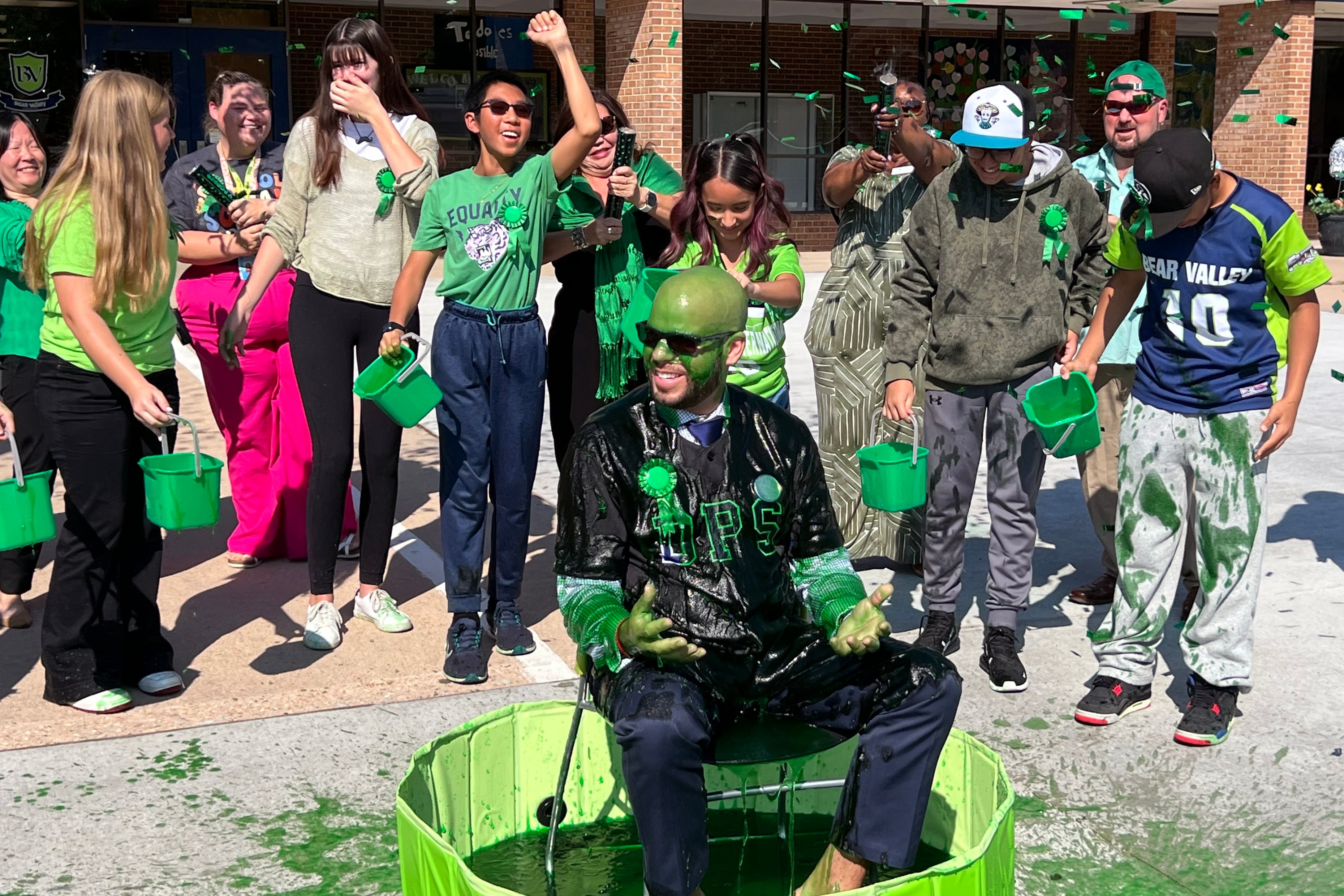 A photograph of a man sitting in a blue tub with green pain on him while a group of young students stand behind him cheering. There is a large school in the background.