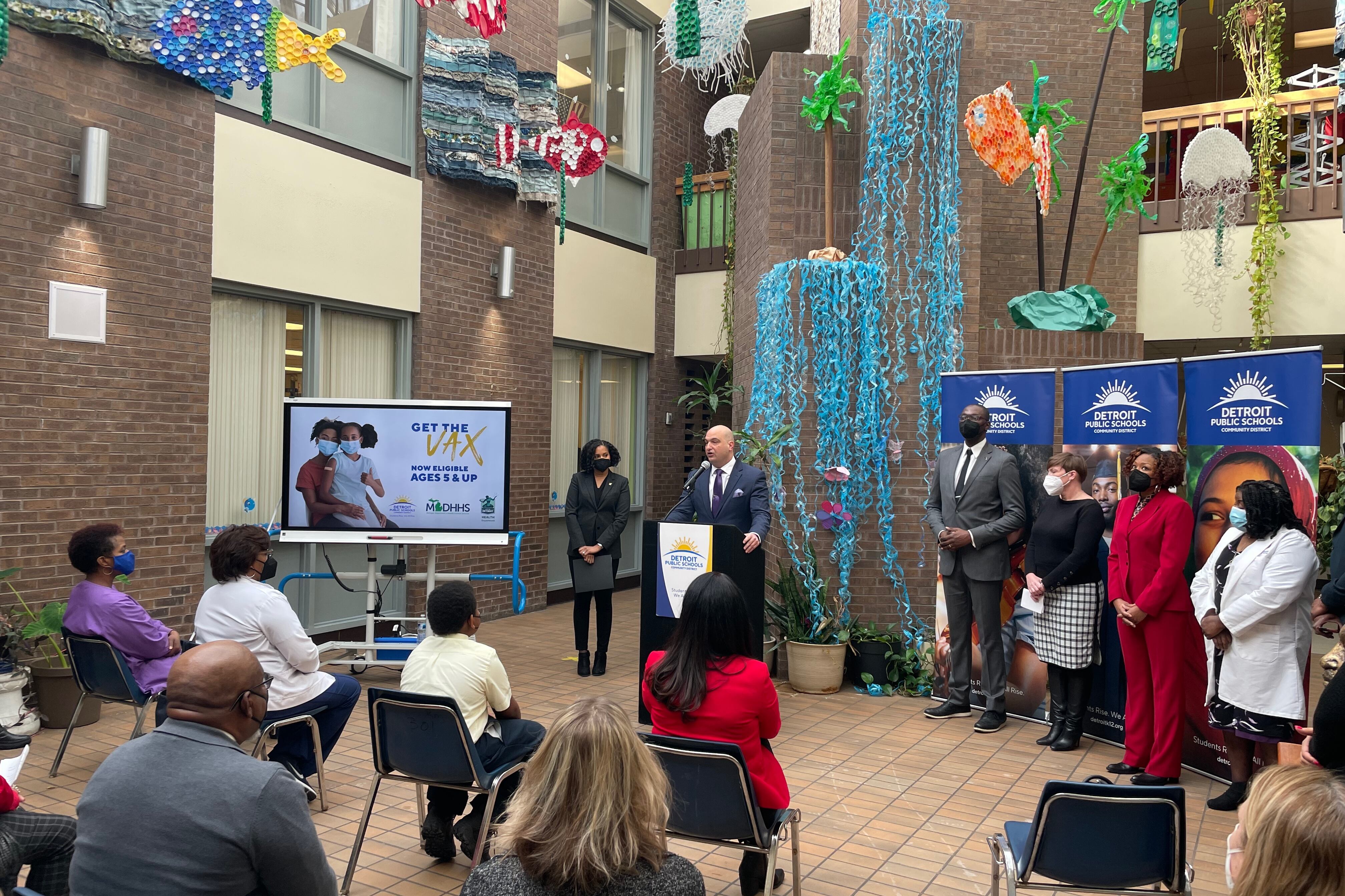 School superintendent speaks to a sitting crowd and across from a TV monitor projecting an image about the COVID vaccine. Four other speakers stand to the right of the superintendent.