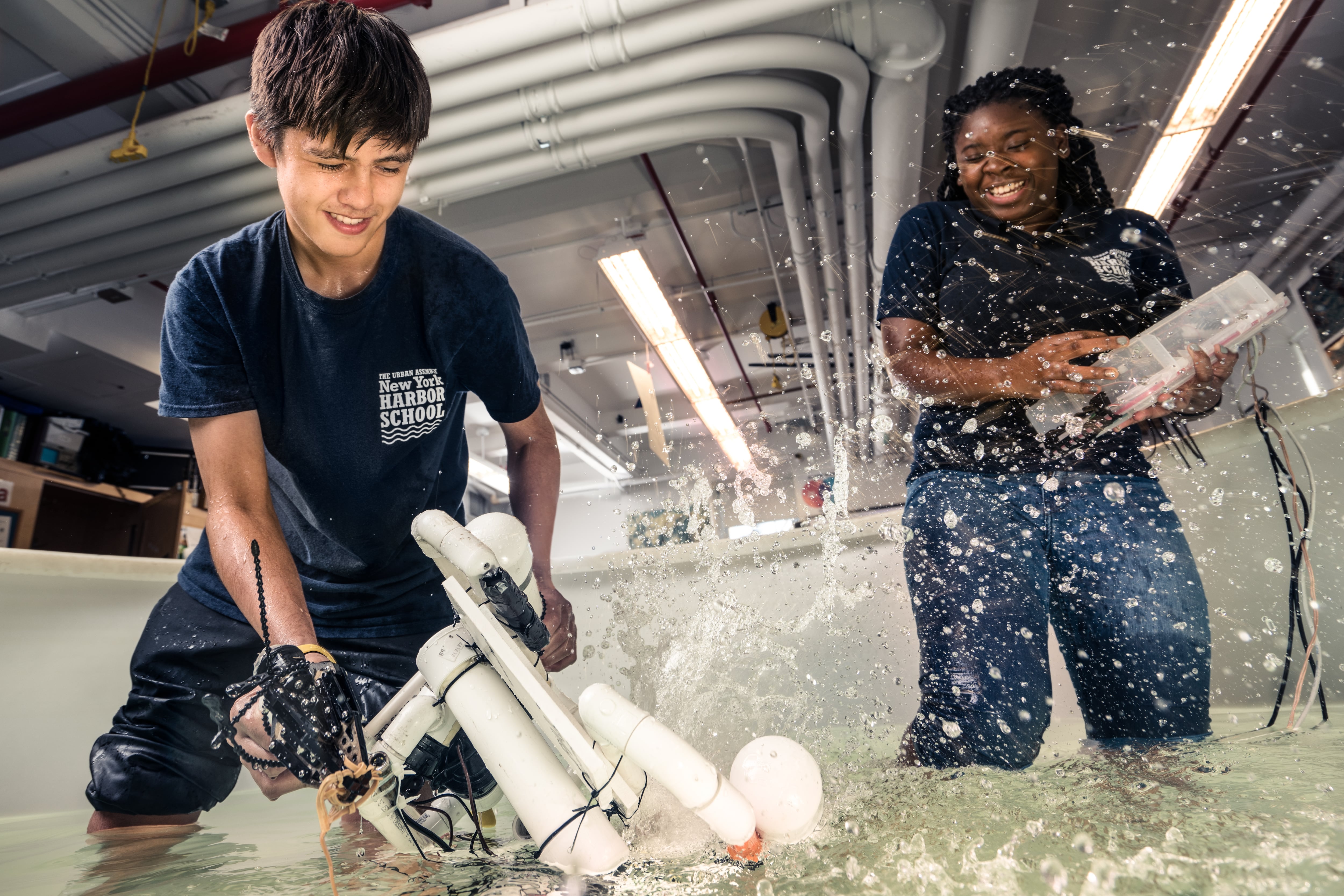 Two students wearing navy blue Urban Assembly New York Harbor School T-shirts get splashed as they play with robotic contraptions.