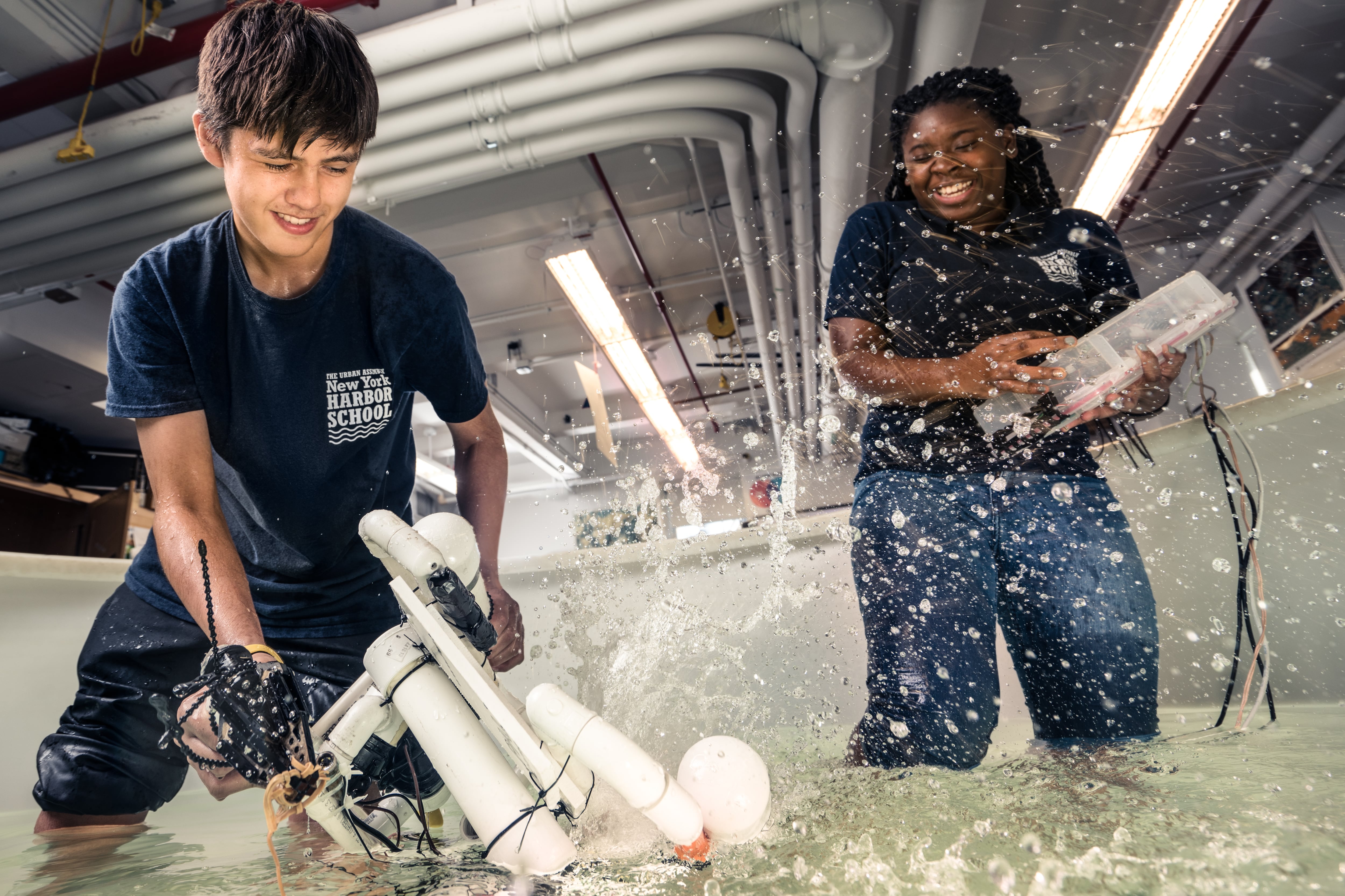 Two students wearing navy blue Urban Assembly New York Harbor School T-shirts get splashed as they play with robotic contraptions.
