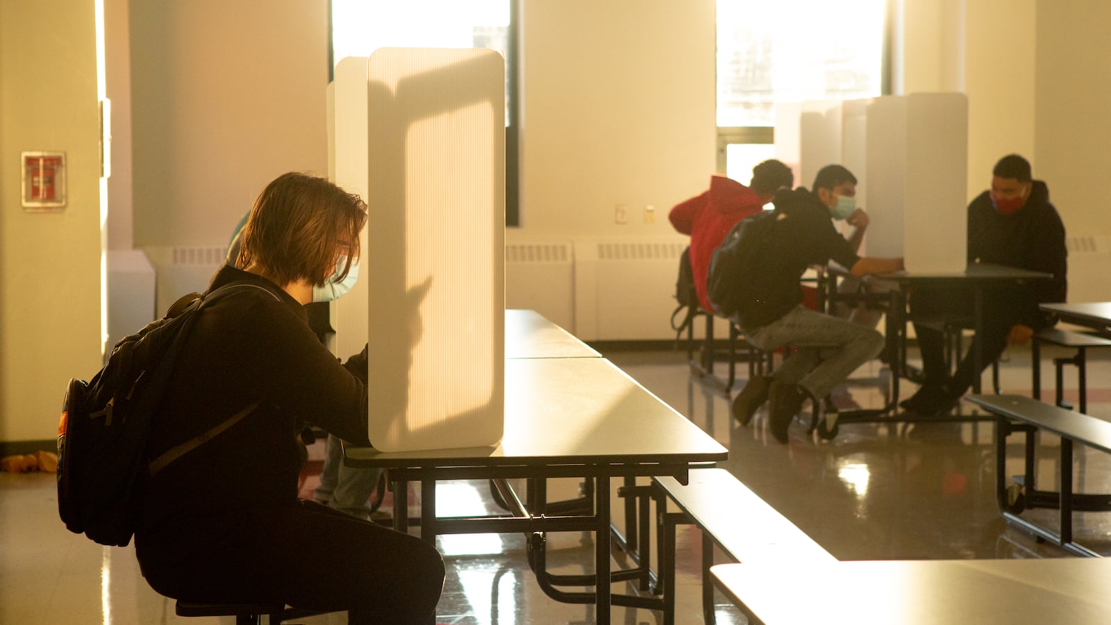 A student wearing a backpack sits on a bench at a table with a trifold partition on top, while three male students in the background also sit at table with partitions.