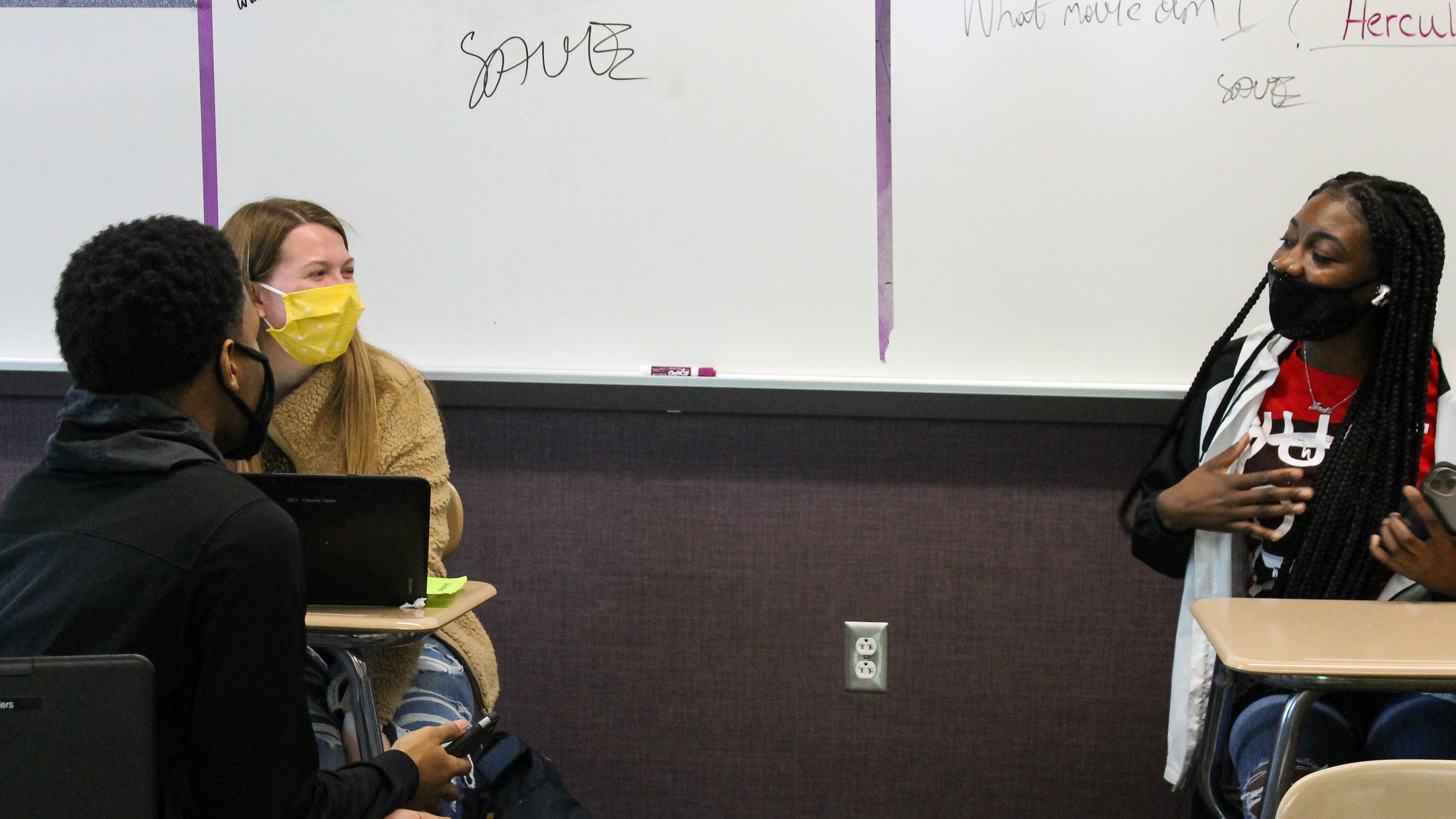 In front of a classroom white board, a student in a yellow sweater wearing a bright yellow mask sits next to a student in a black sweatshirt with a black mask. They are talking to another student seated in a desk several feet away with a red shirt and a black face mask holding her hand to her chest in the middle of speaking.