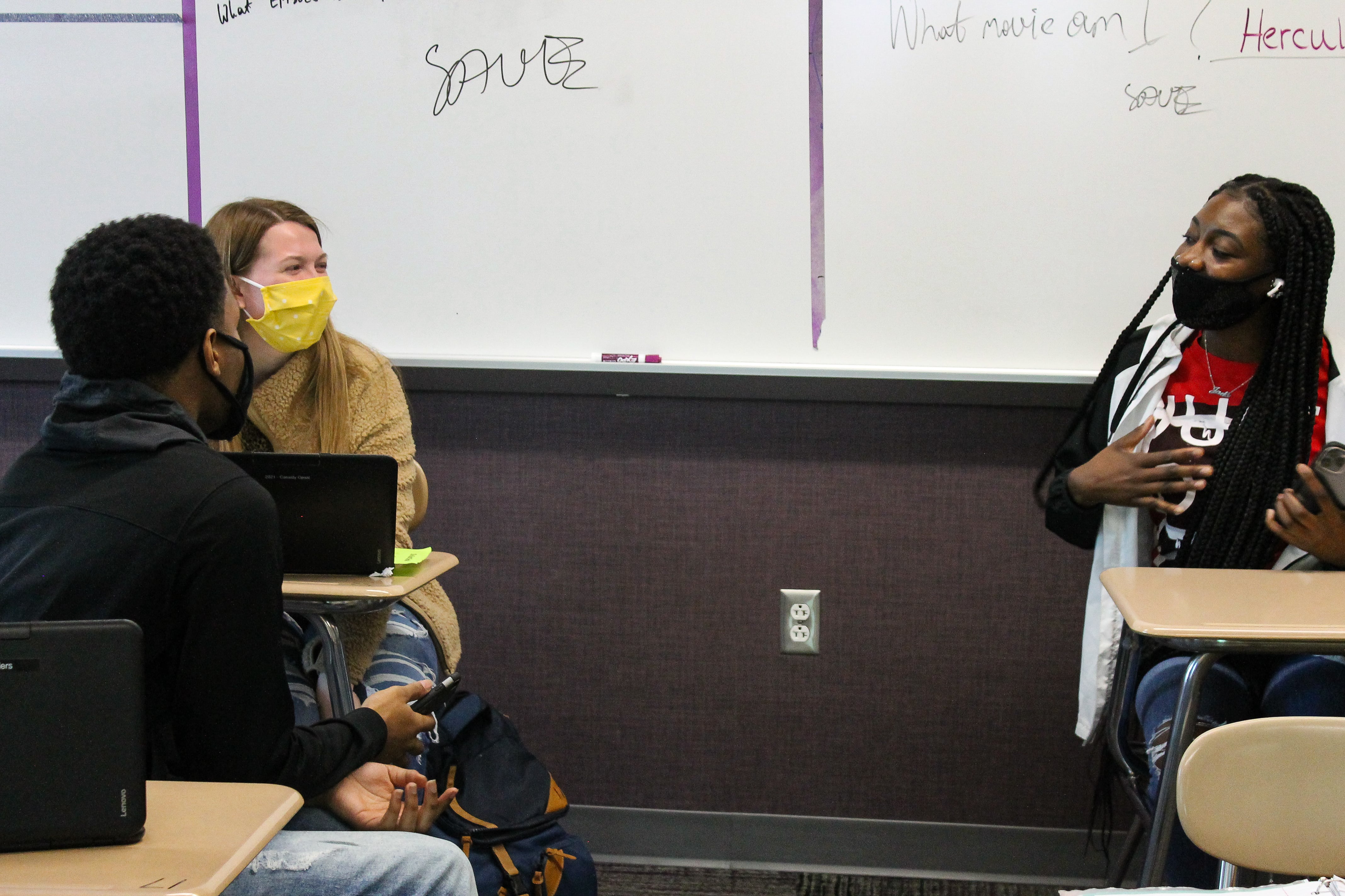 In front of a classroom white board, a student in a yellow sweater wearing a bright yellow mask sits next to a student in a black sweatshirt with a black mask. They are talking to another student seated in a desk several feet away with a red shirt and a black face mask holding her hand to her chest in the middle of speaking.