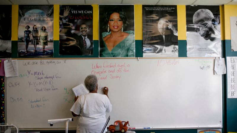 A teacher writes on a dry-erase board in a classroom. Math equations are written on the board to her left. Above the board are posters of Barack Obama, Oprah Winfrey, Martin Luther King Jr., Mahatma Gandhi, and the cast of the movie Hidden Figures.