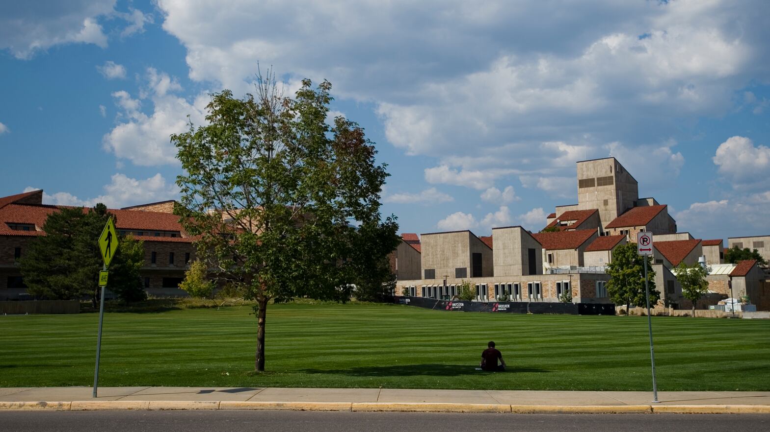 A University of Colorado Boulder student studies in the shade of a tree in front of Business Field with school buildings in the background.