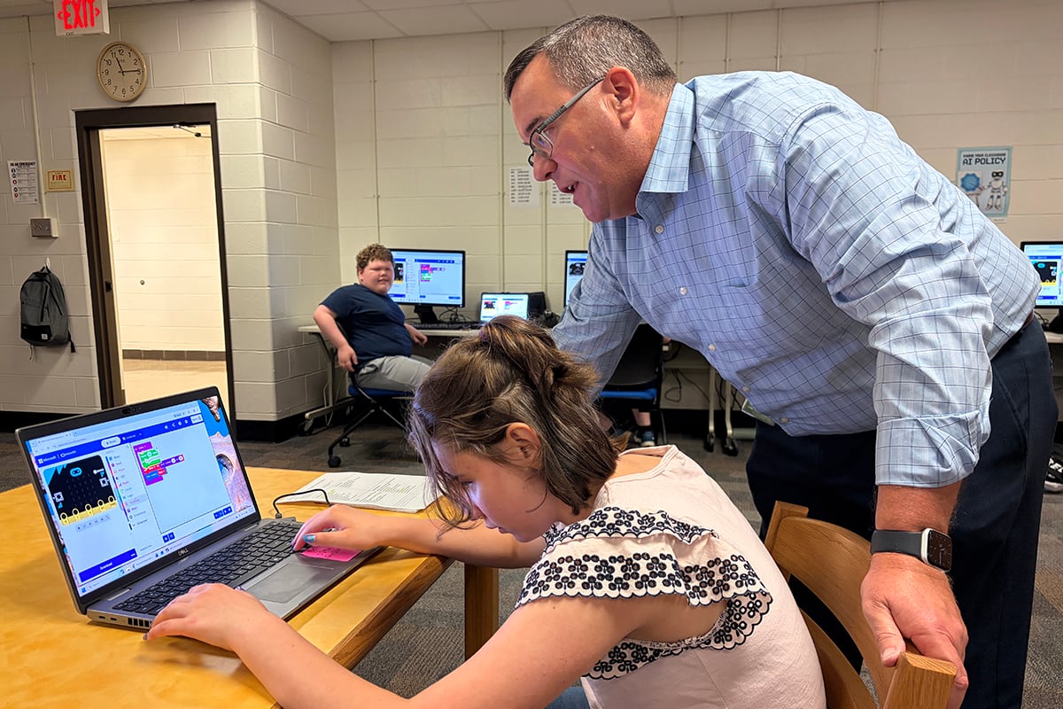 A girl in a light-color top works on her computer as her teacher in a blue shirt watches.