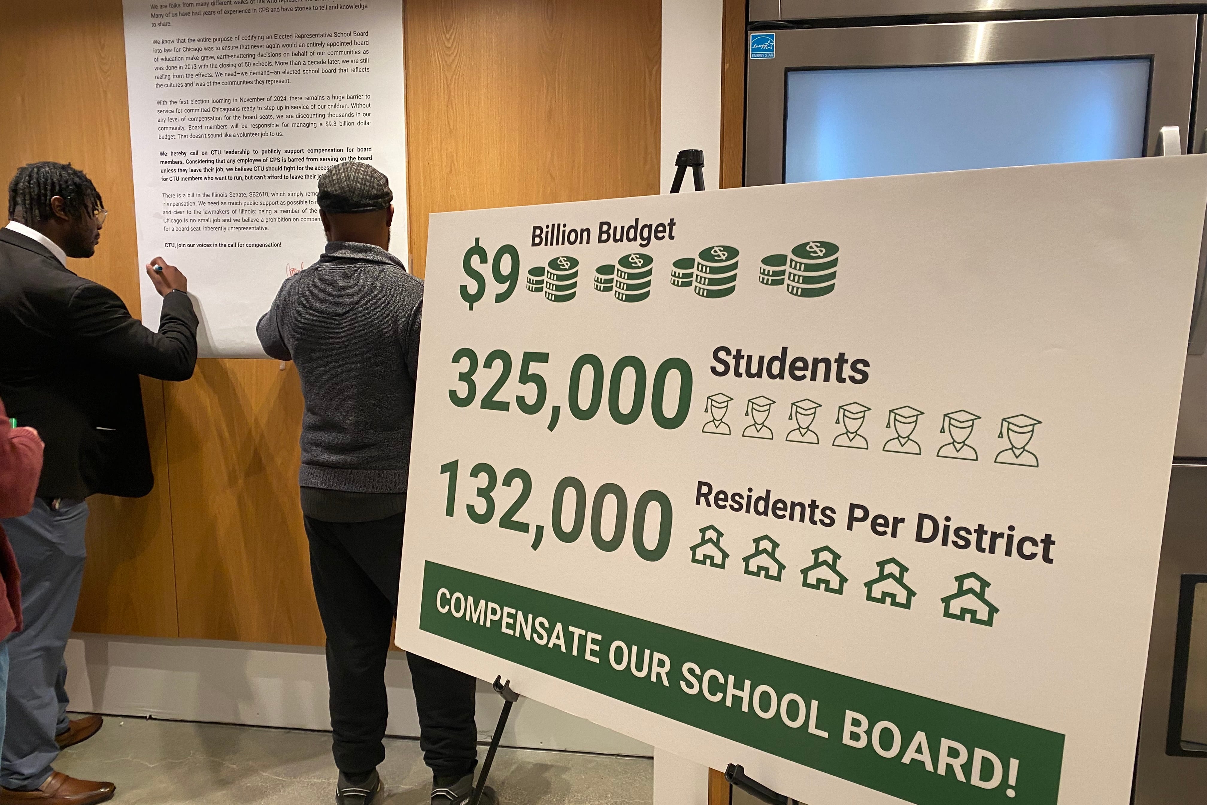 A sign urging for Chicago school board members to be compensated in the forefront. People sign a letter on a wall in the background.