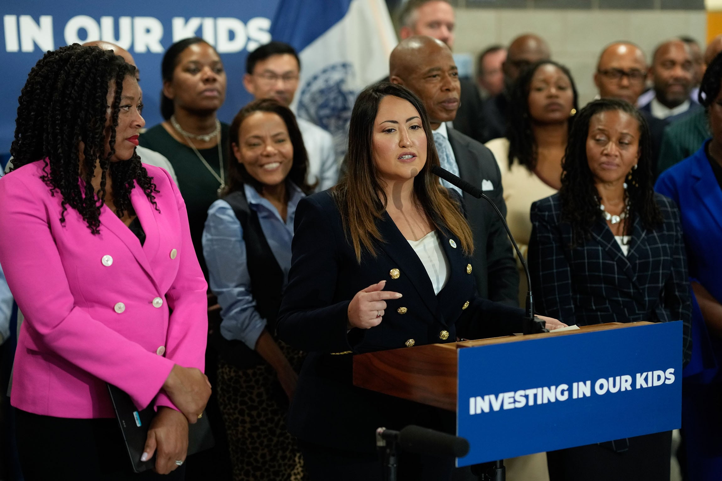 A woman in a business suit speaks from behind a podium with a large group of people behind her.