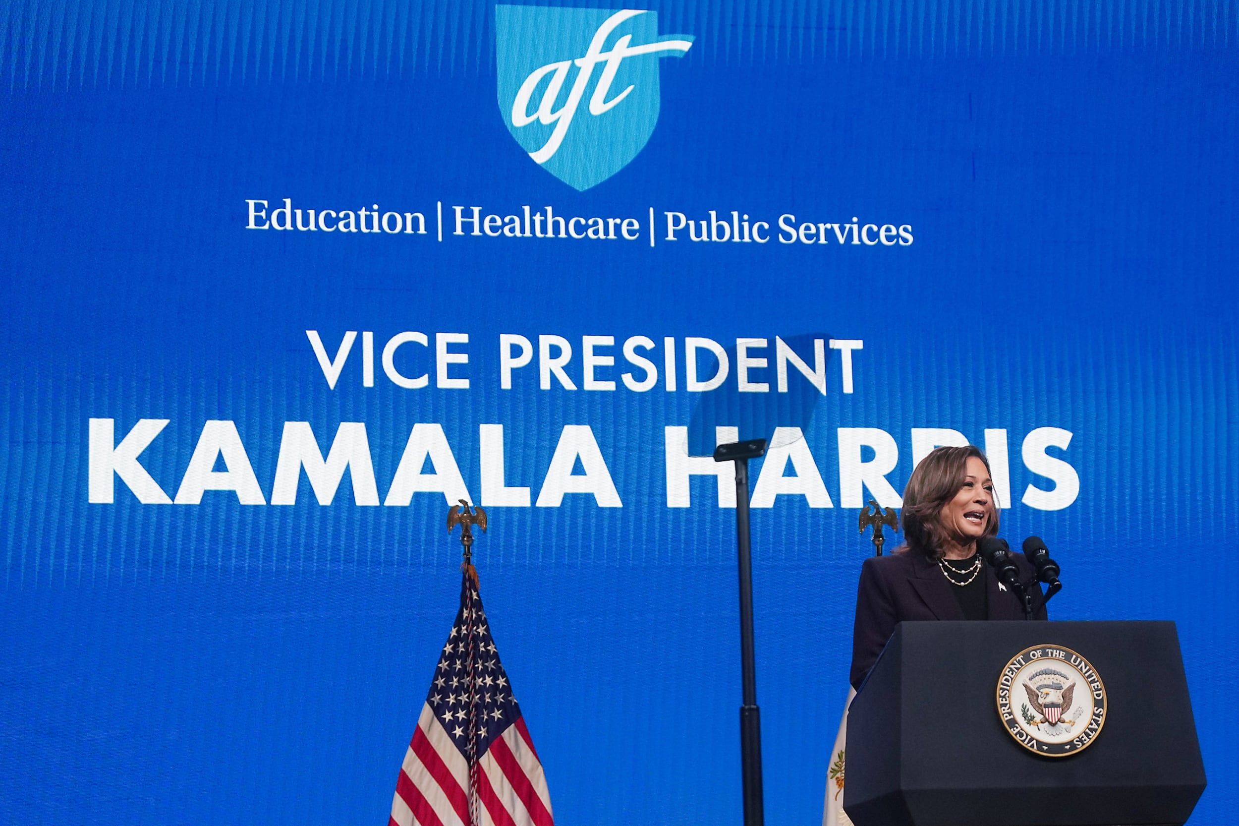 A woman with dark hair and wearing a dark suit stands behind a podium with a microphone with a blue screen in the background.