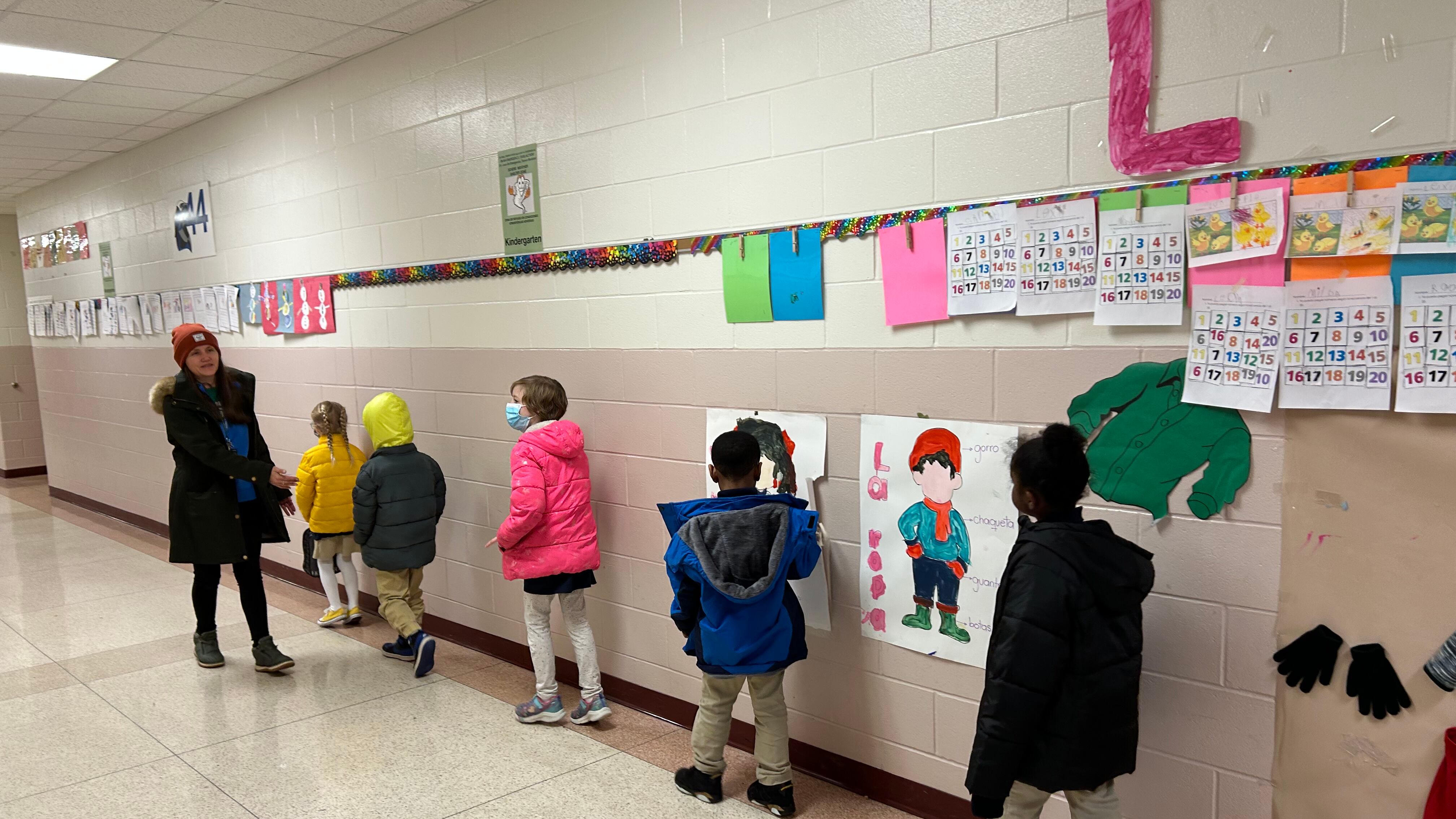 Five students walk in a school hallway beside a teacher who looks back and guides them.