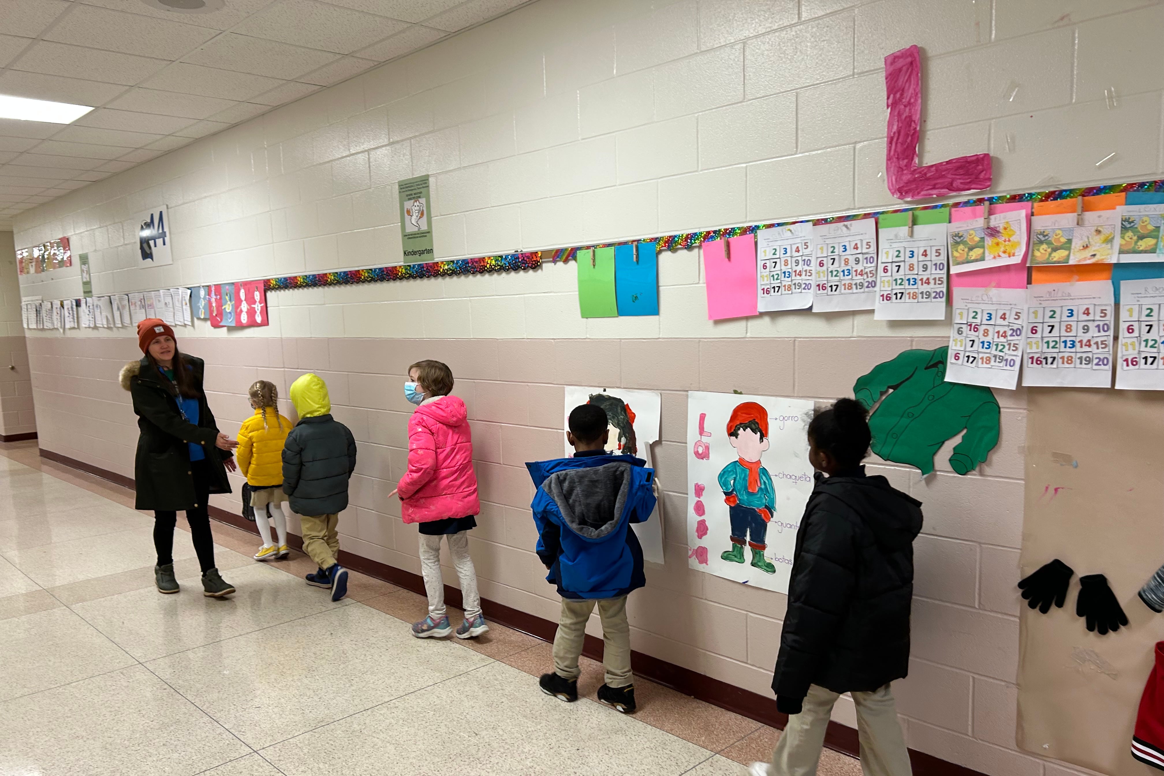 Five students walk in a school hallway beside a teacher who looks back and guides them.