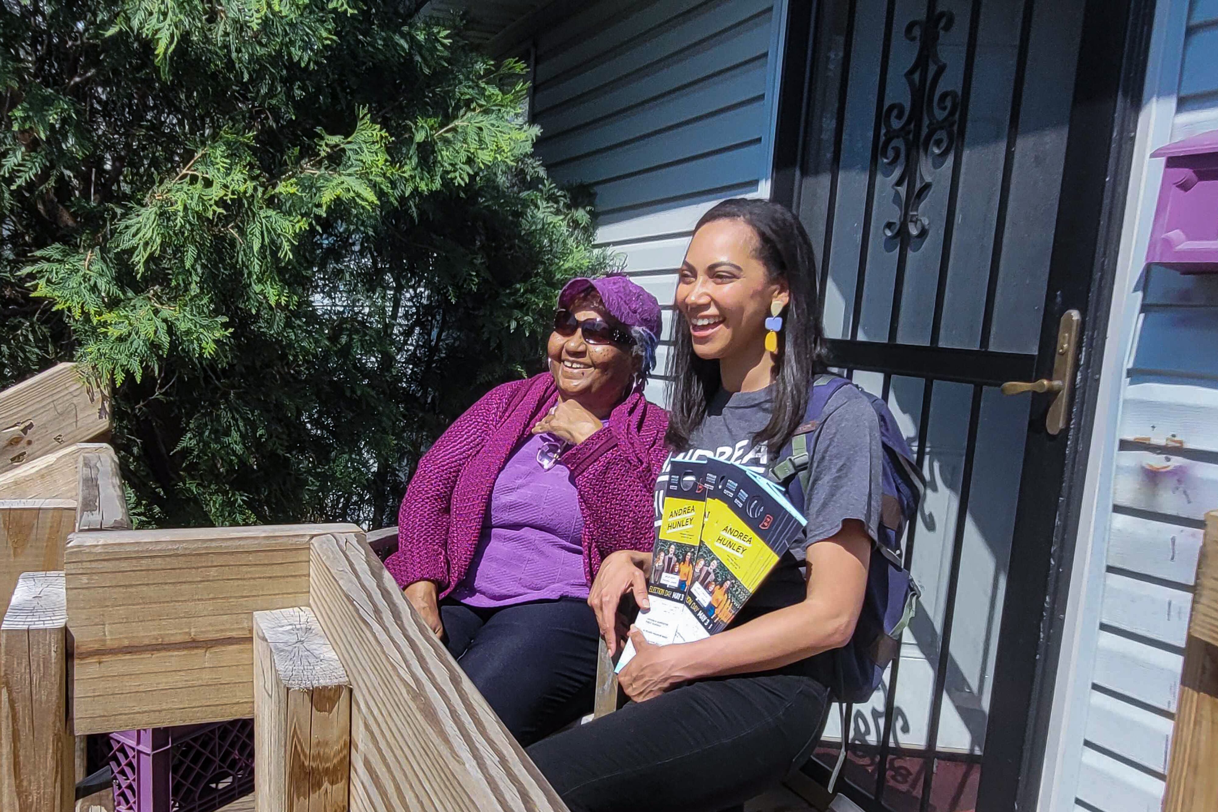 Two women, one in a purple sweater and purple shirt and hat, and black pants, the other wearing a gray campaign T-shirt and carrying fliers, sit smiling on the porch of a home.