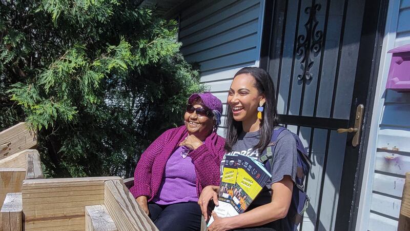 Two women, one in a purple sweater and purple shirt and hat, and black pants, the other wearing a gray campaign T-shirt and carrying fliers, sit smiling on the porch of a home.
