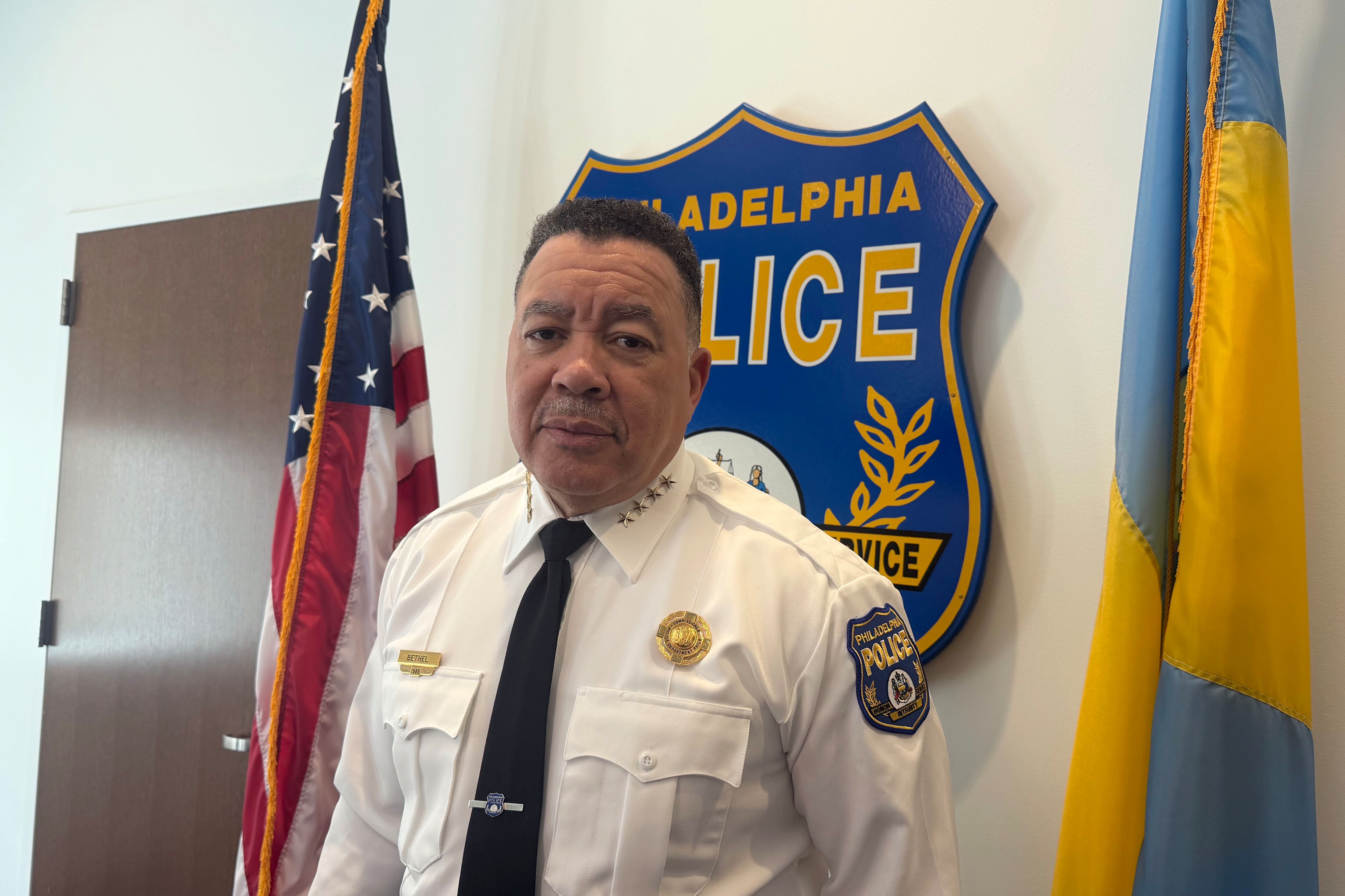 A photograph of a Black man in a police commissioner uniform poses for a portrait in front of a large police emblem and two flags.