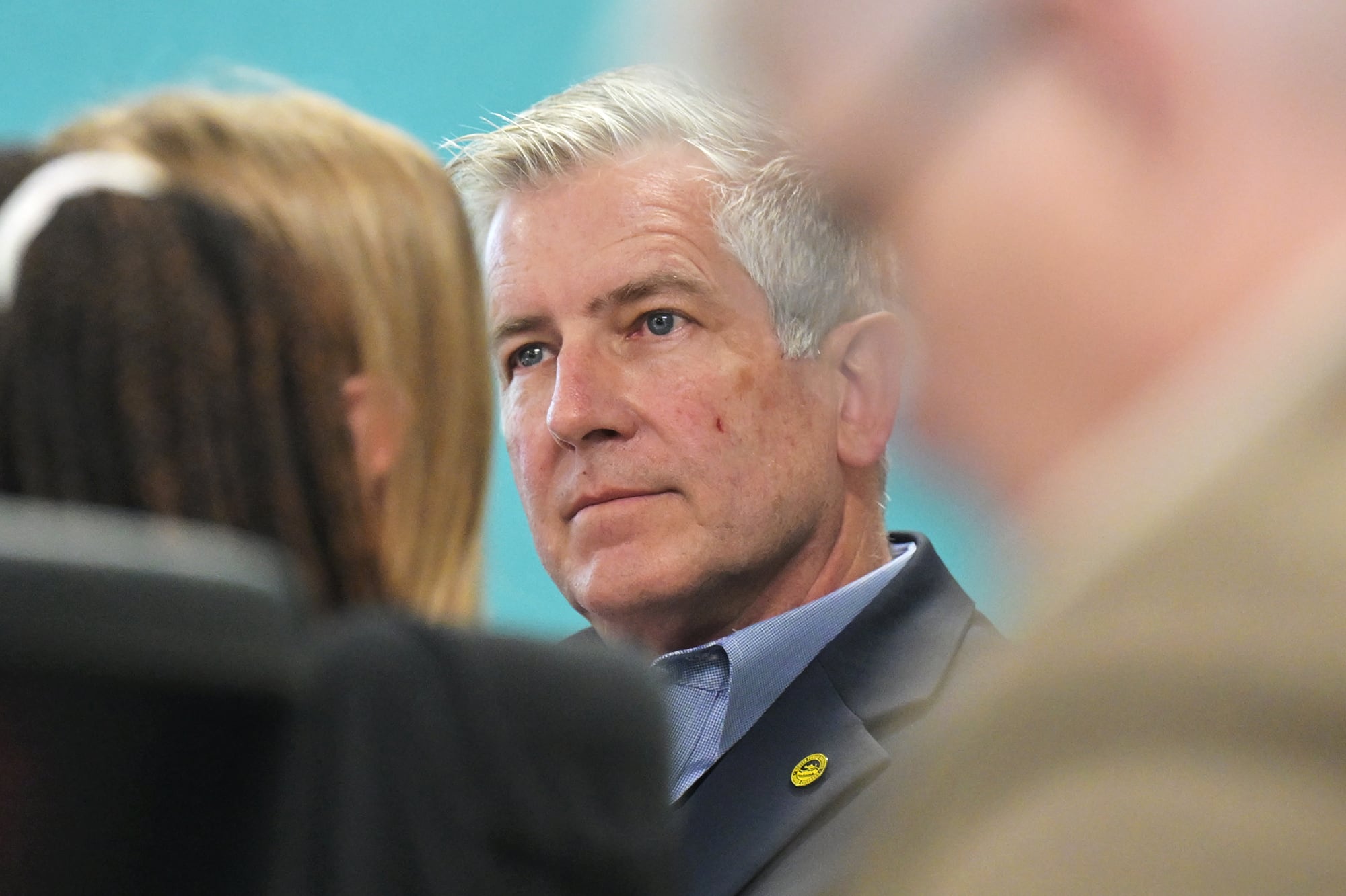 A photograph of a white man in a suit sitting in a room seen through a group of people.