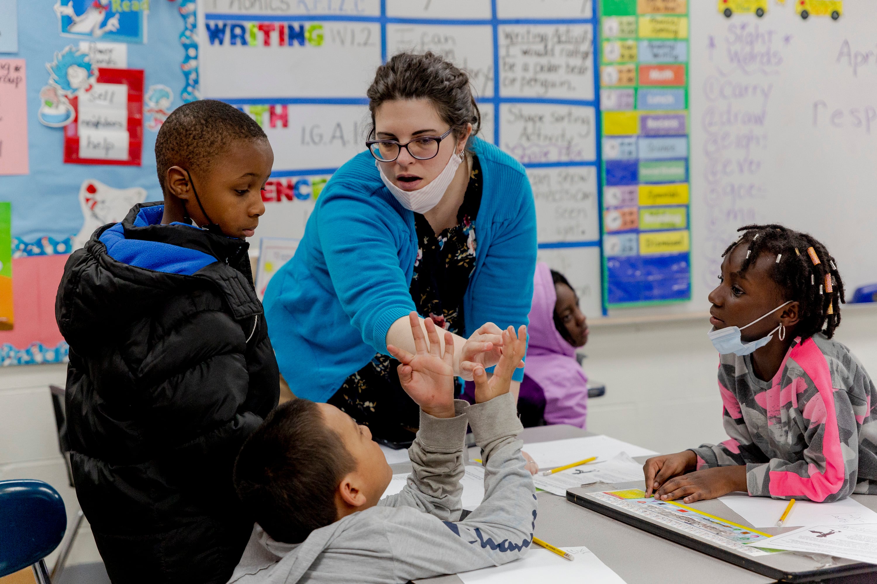 A teacher wearing a blue shirt helps her student count numbers during a math lesson in her classroom, using his fingers to figure out a problem.