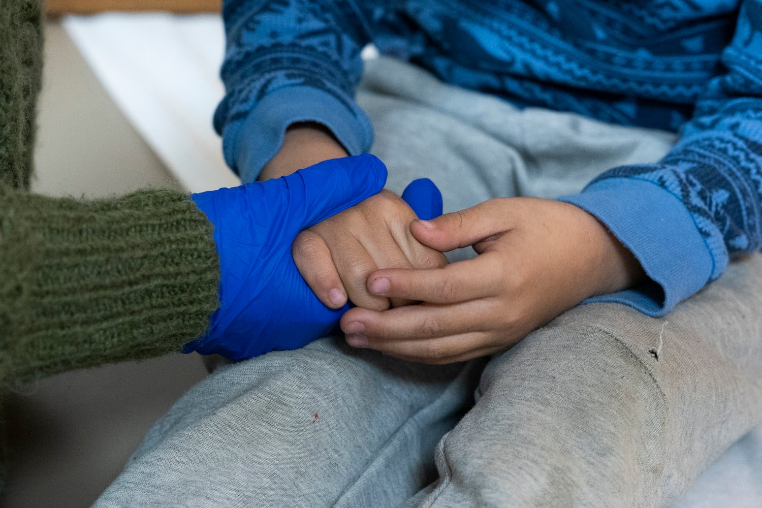 A photograph up close up a medical professional wearing blue medical gloves and holding the hands of a child.