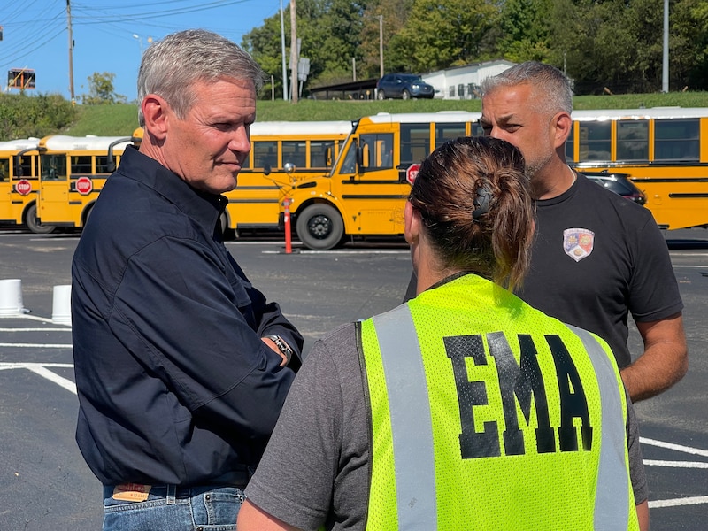 Three people stand outside in front of several yellow and black school buses.