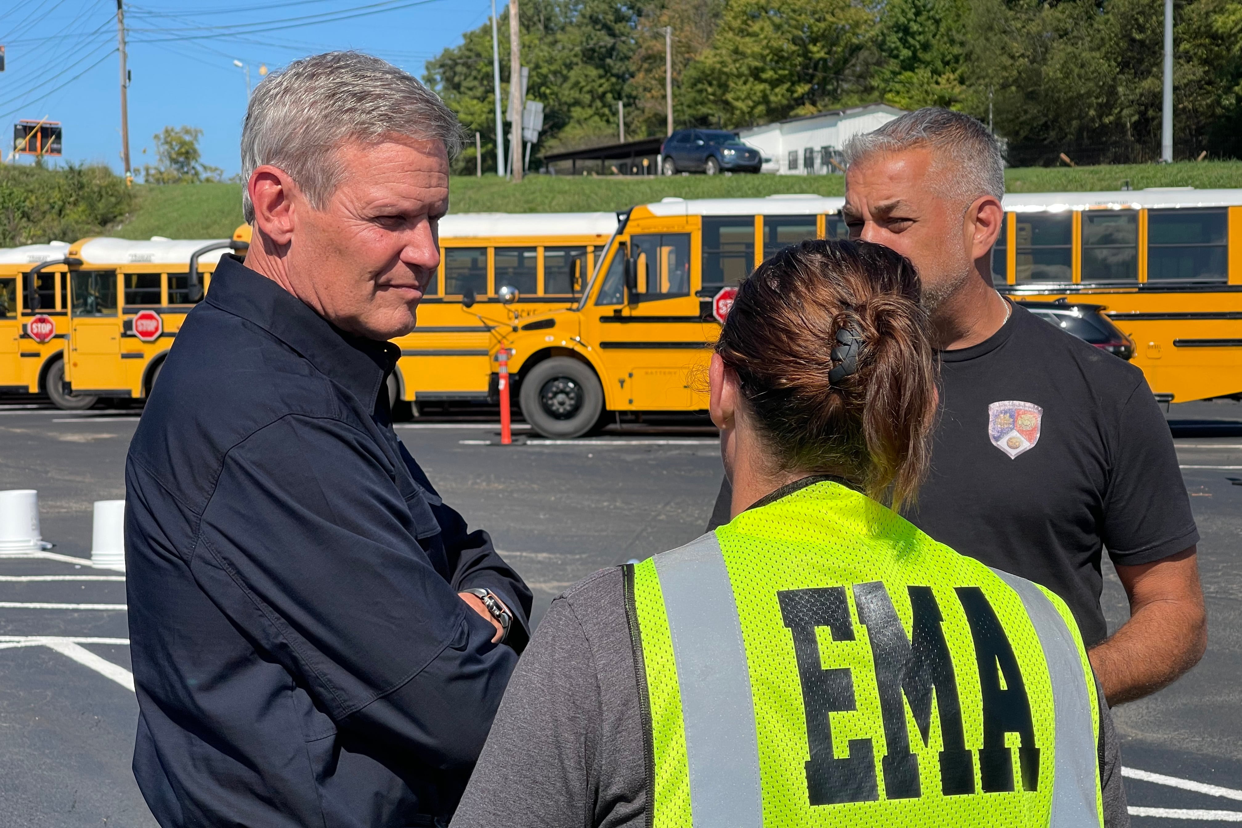 Three people stand outside in front of several yellow and black school buses.