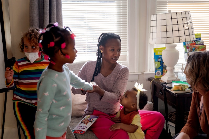 An adult woman wearing a purple shirt sits in a chair with three children standing around her and another adult in a house near a large window.