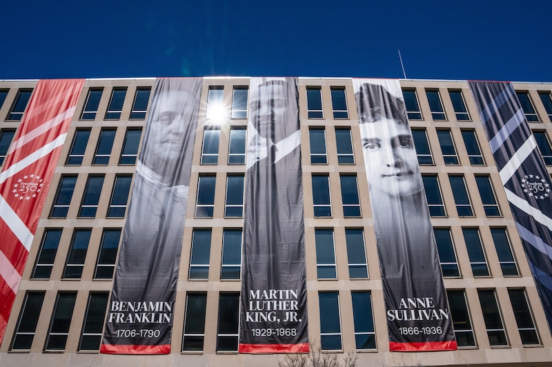 Large vertical banners hang from a brutalist office building. They depict Benjamin Franklin, Martin Luther King Jr., and Anne Sullivan.