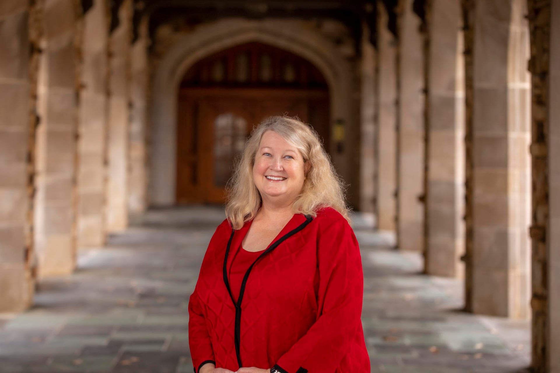 A women with blonde hair poses for a portrait in front of two large rows of columns. She is reading a bright red blouse with black trim.