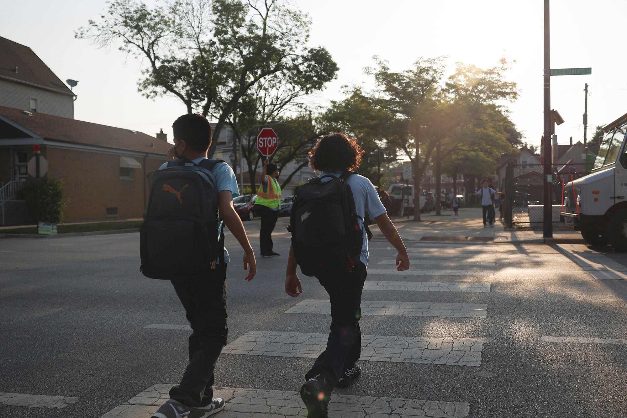 A photograph of two young students wearing backpacks cross the street early in the morning.