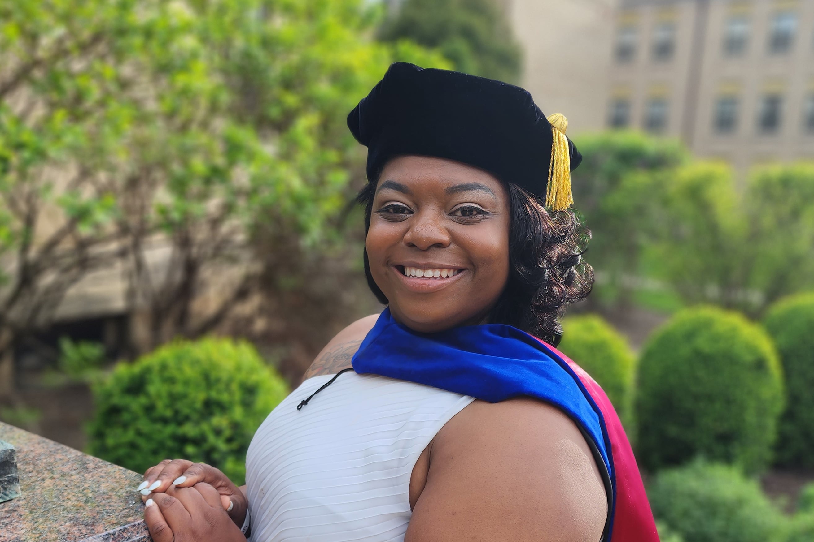 A woman wearing a white dress and a graduation cap and honors sash smiles for a portrait outside with green trees in the background.