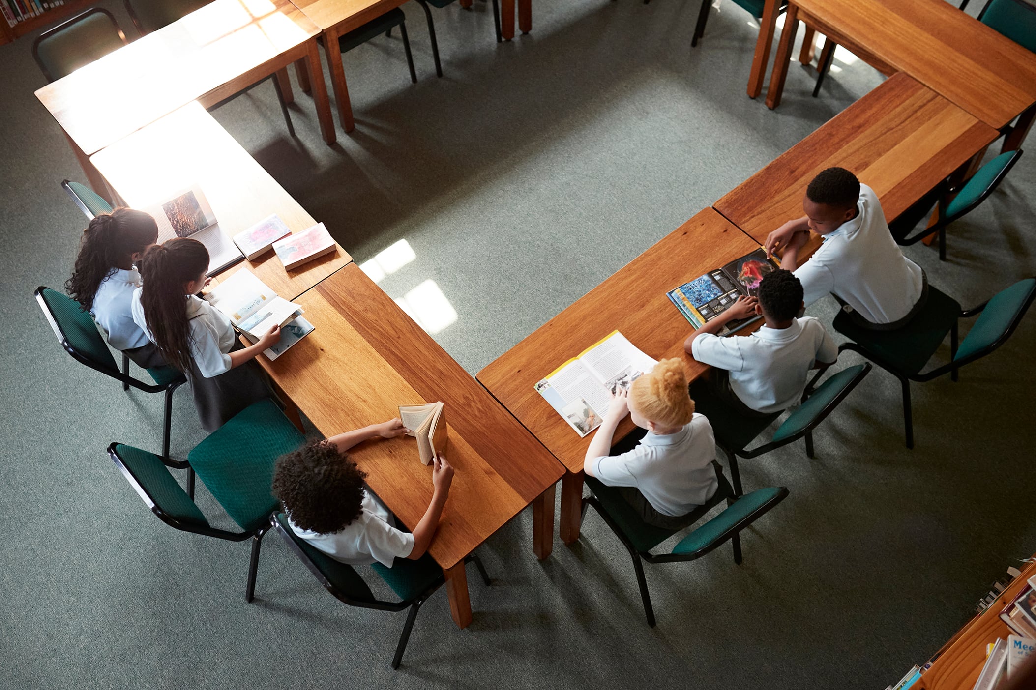 An aerial photograph of six middle schoolers all wearing the same light blue shirt and uniform sitting around a wooden table in a library.