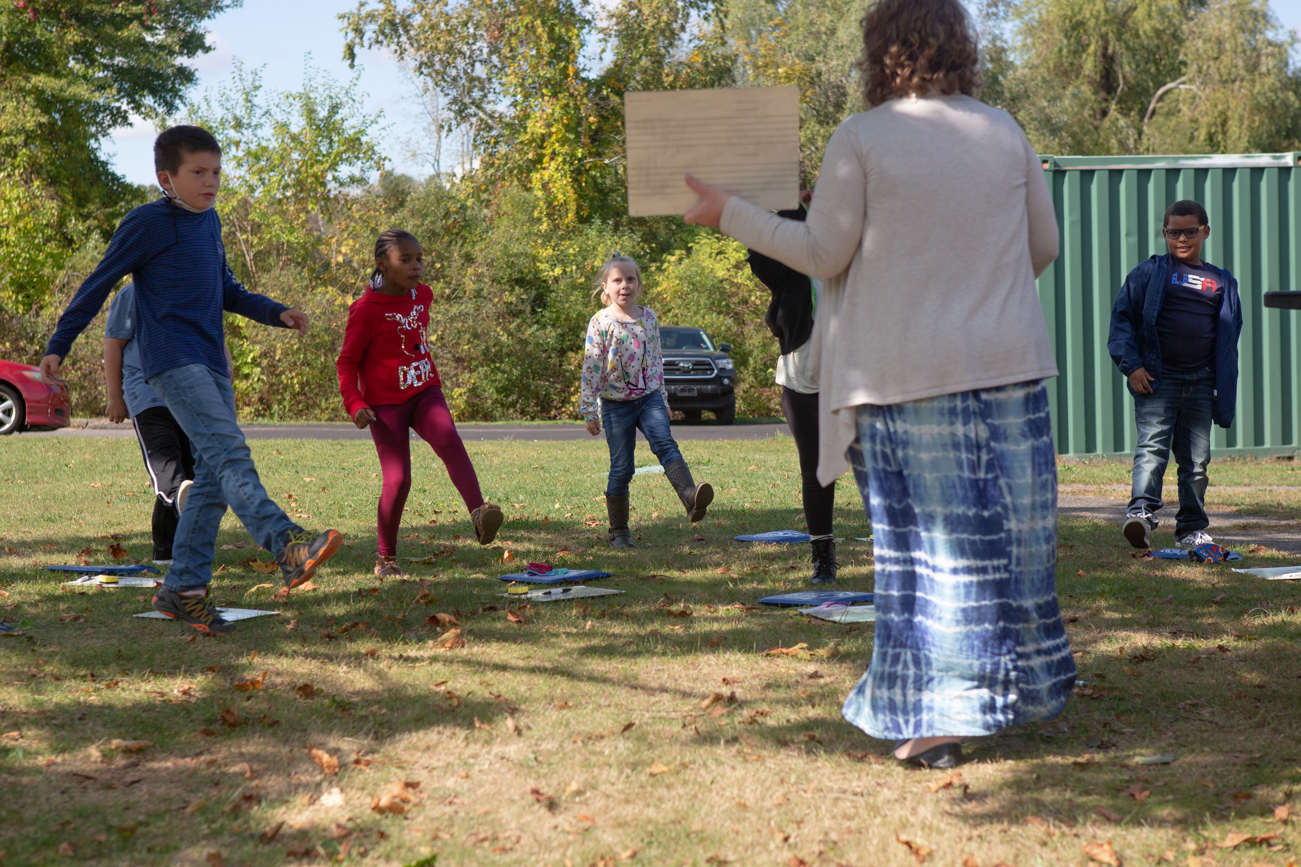 A teacher in a white sweater and a long blue-and-white skirt stand on a lawn facing about a half-dozen standing students who are each lifting one leg together.