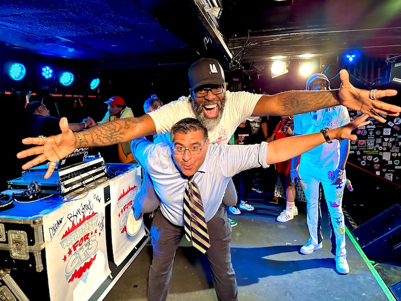 A man in a button-down shirt and tie gives another man wearing a T-shirt and baseball cap a piggyback ride near DJ equipment.