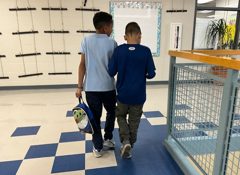 Two young boys wearing blue shirts walk down a school hallway.