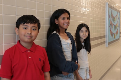 Three young middle school students pose for a portrait in a school hallway.