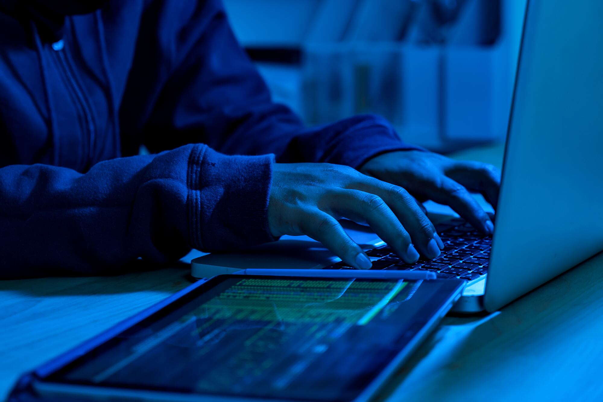 With blue light cast on the photo, a pair of hands type on a laptop. A tablet is seen in the foreground.