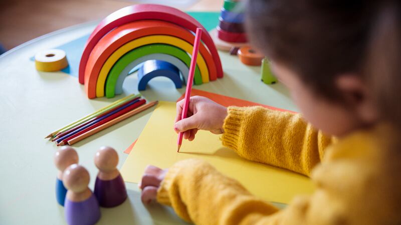 A picture of a young child in a yellow sweater uses a red coloring pencil to draw on a piece of paper.