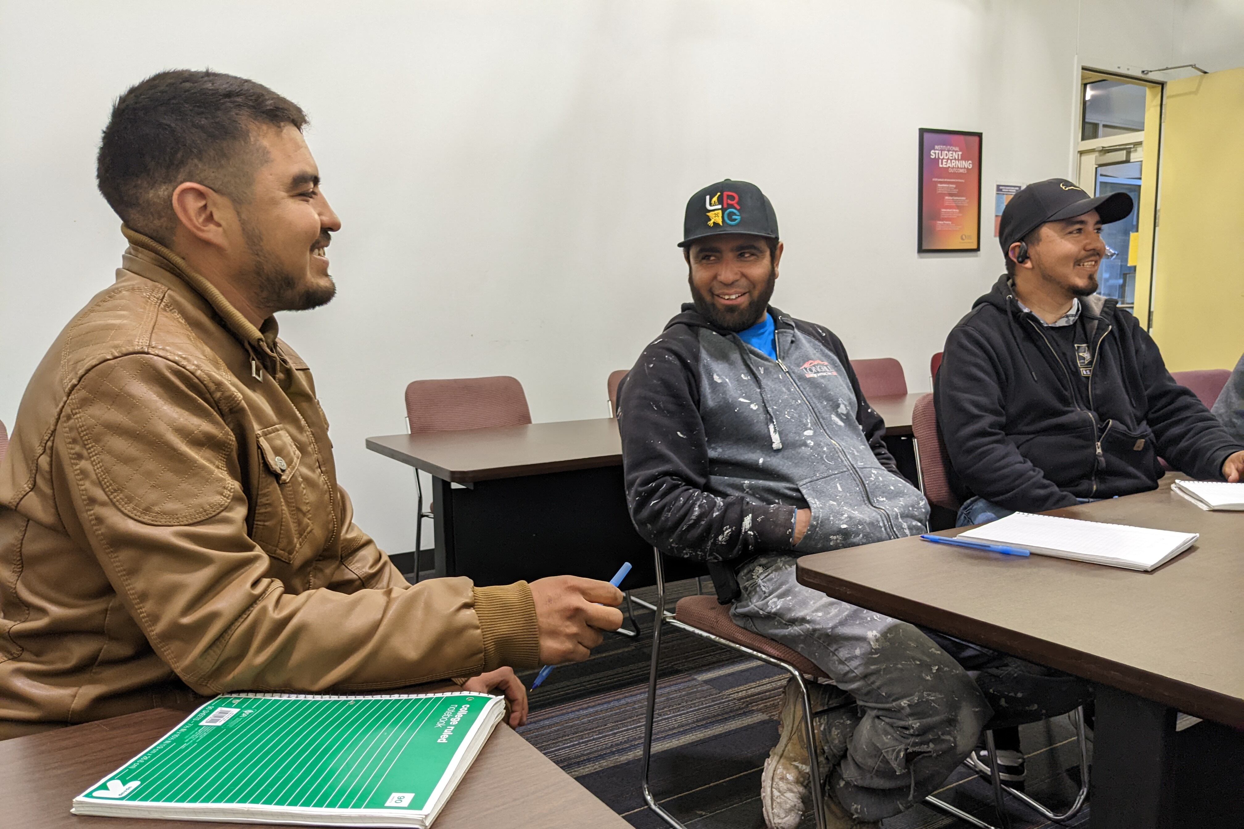 Three men sit at a desk in a classroom.