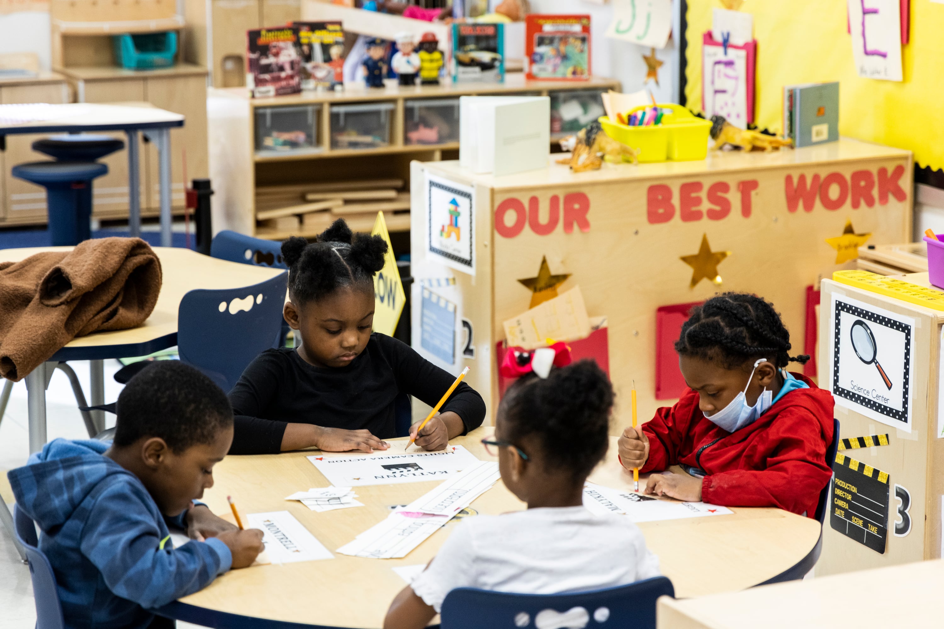 A group of young children sit around a school desk in a classroom.