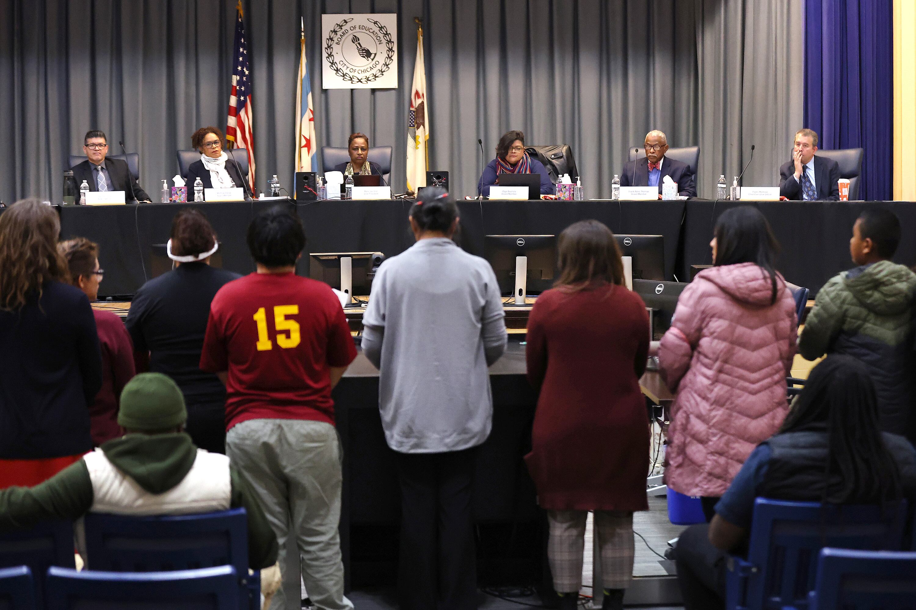 A line of community members face six school board members sitting at a long table each behind a microphone.