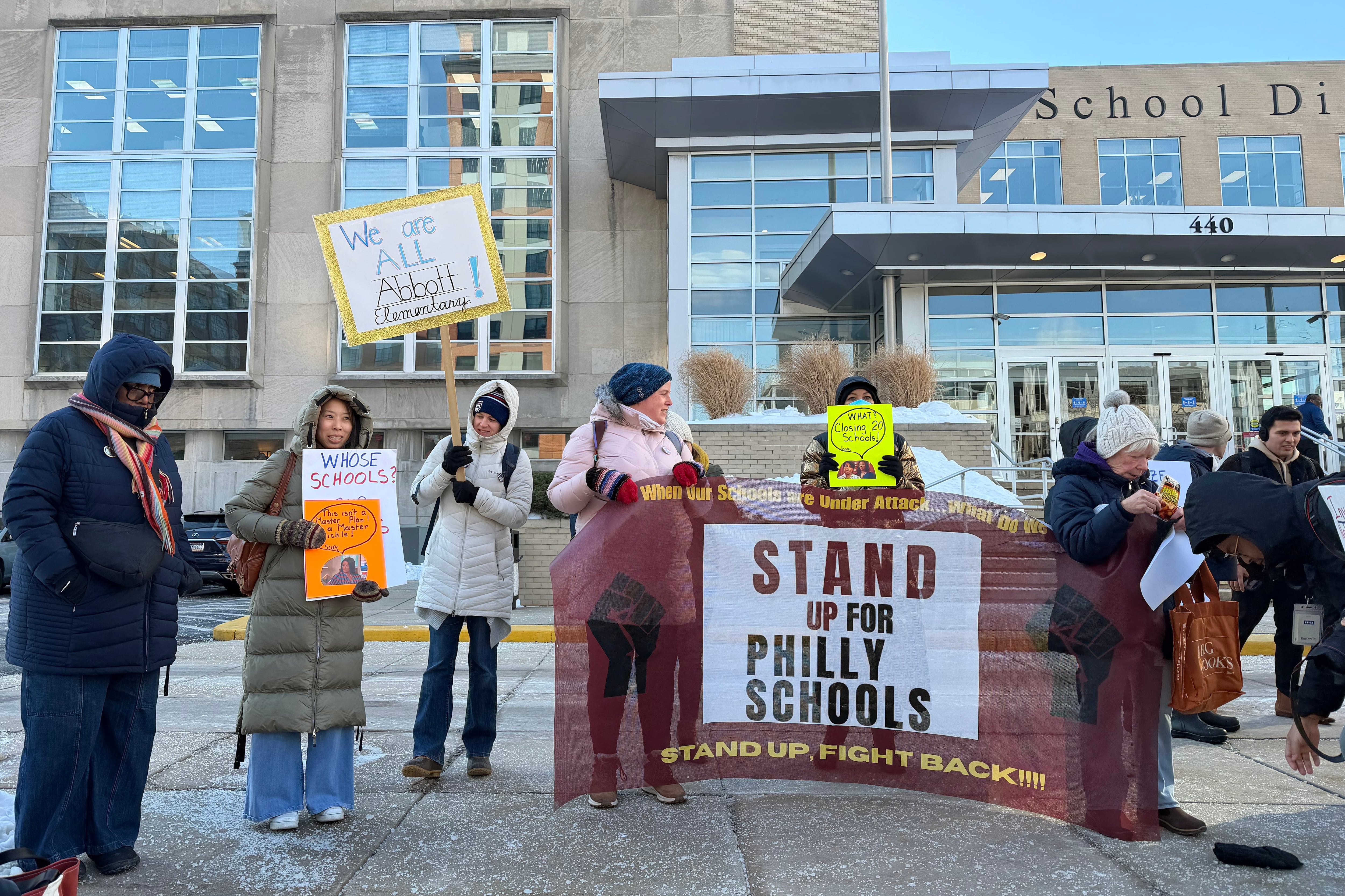 A photograph of a group of people wearing large winter coats and signs protesting outside of a large building.