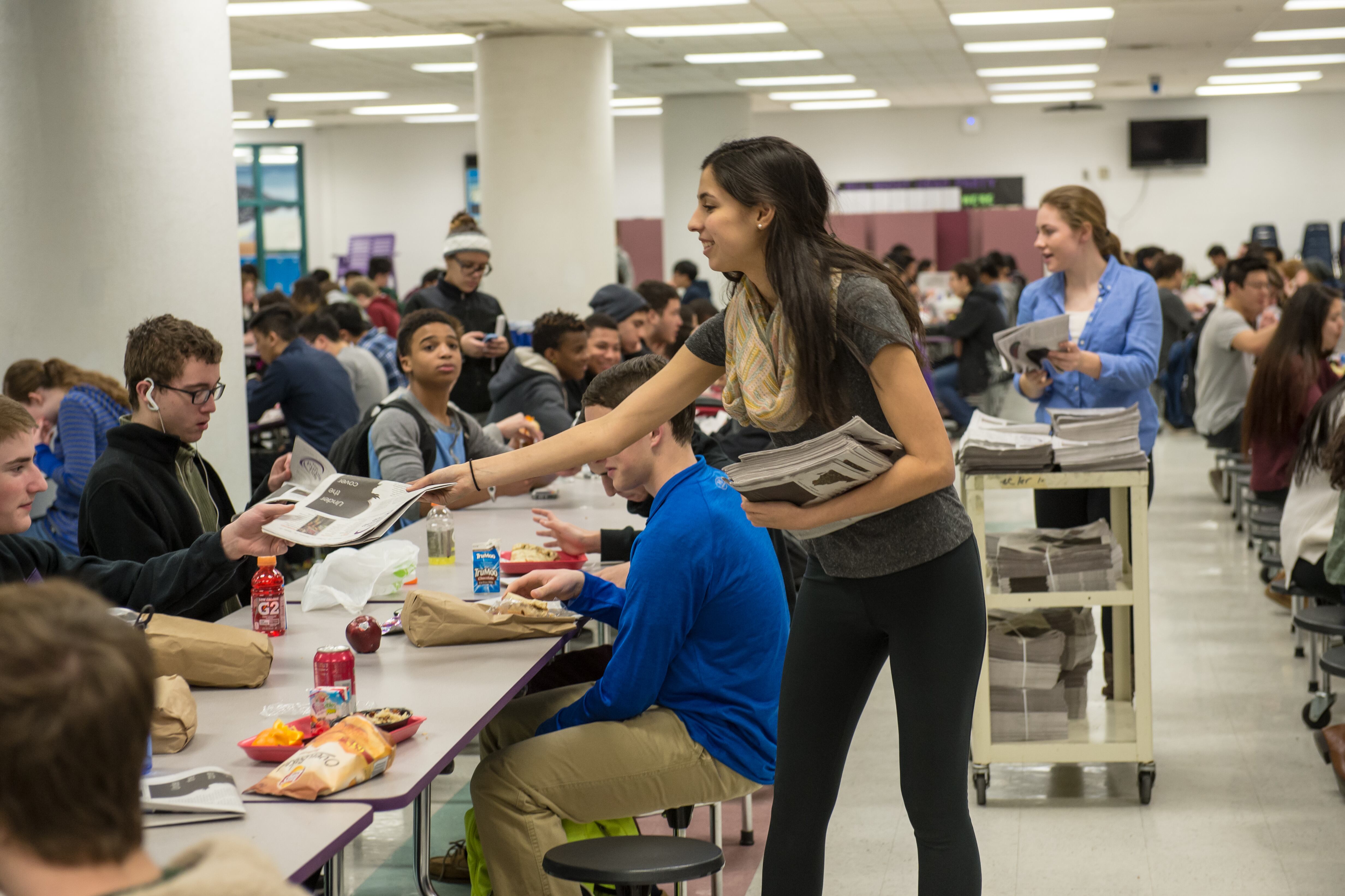 A girl hands out newspapers to students sitting at a lunch table in a school cafeteria.